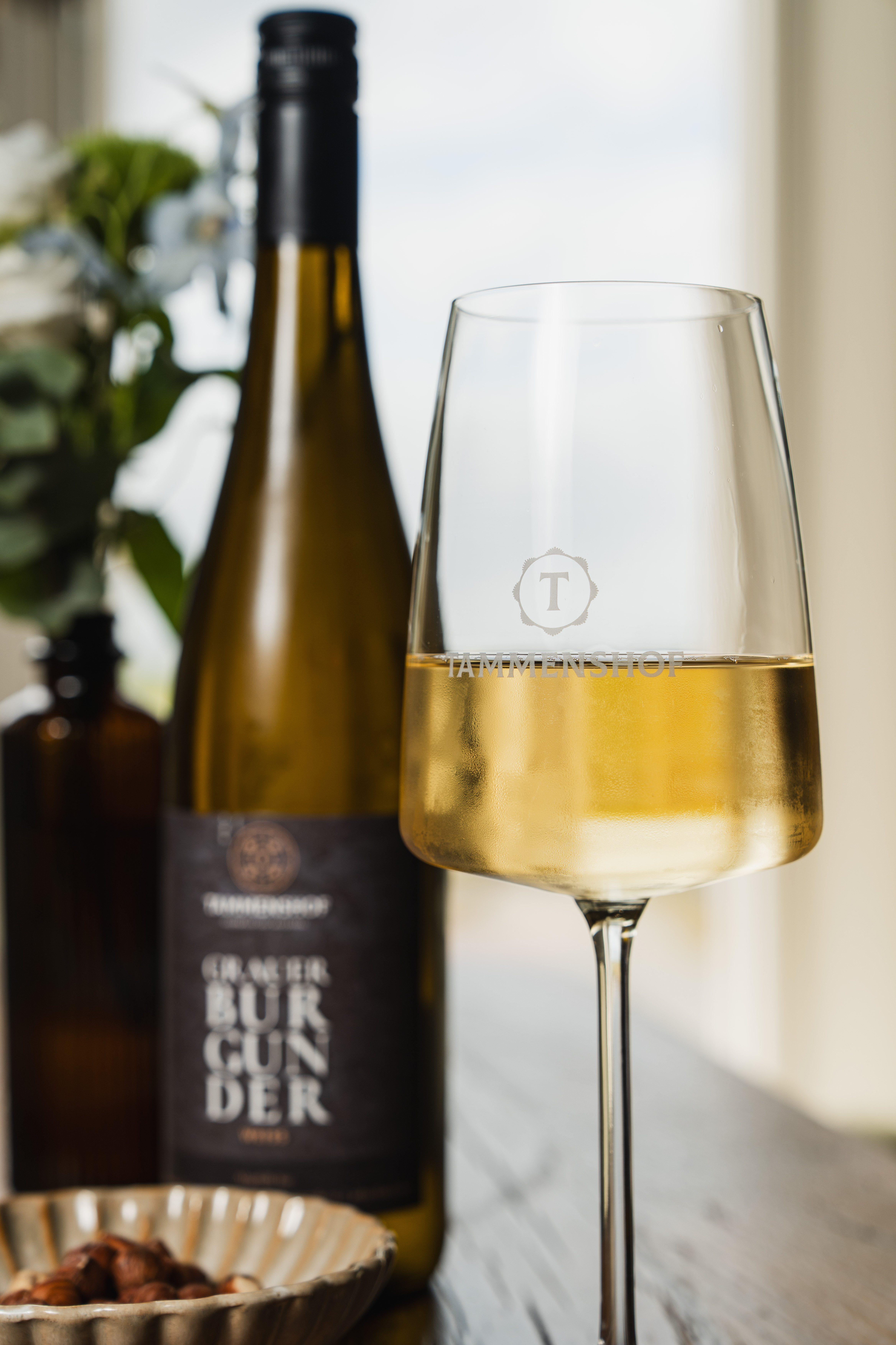 Glass of white wine with a Tammenshof logo beside a wine bottle on a wooden restaurant table.
