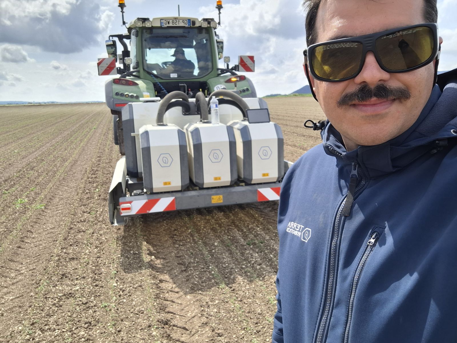 Man with sunglasses and mustache in blue Terra Robotics jacket standing in farmland near tractor equipment.