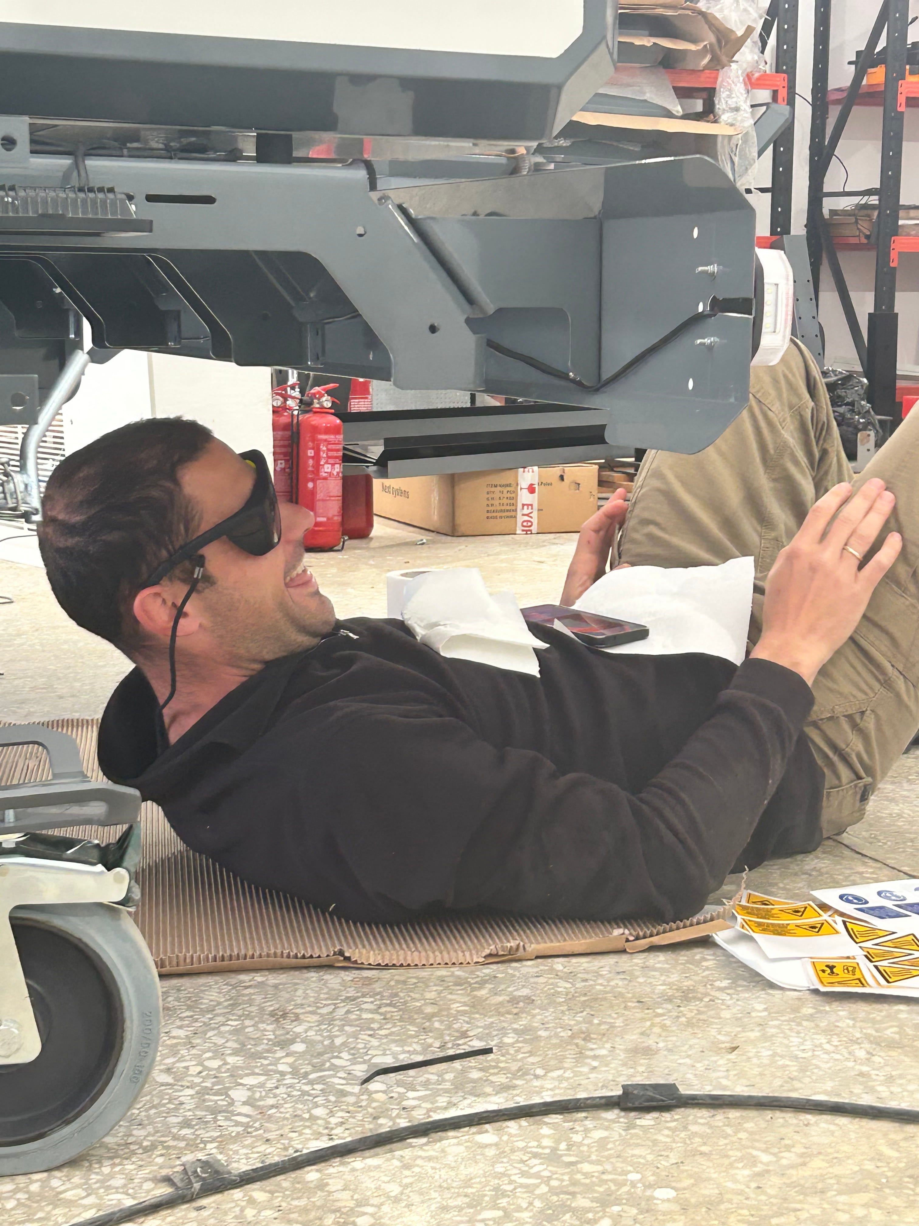 Man wearing sunglasses lies on cardboard under vehicle frame with tools nearby in workshop.