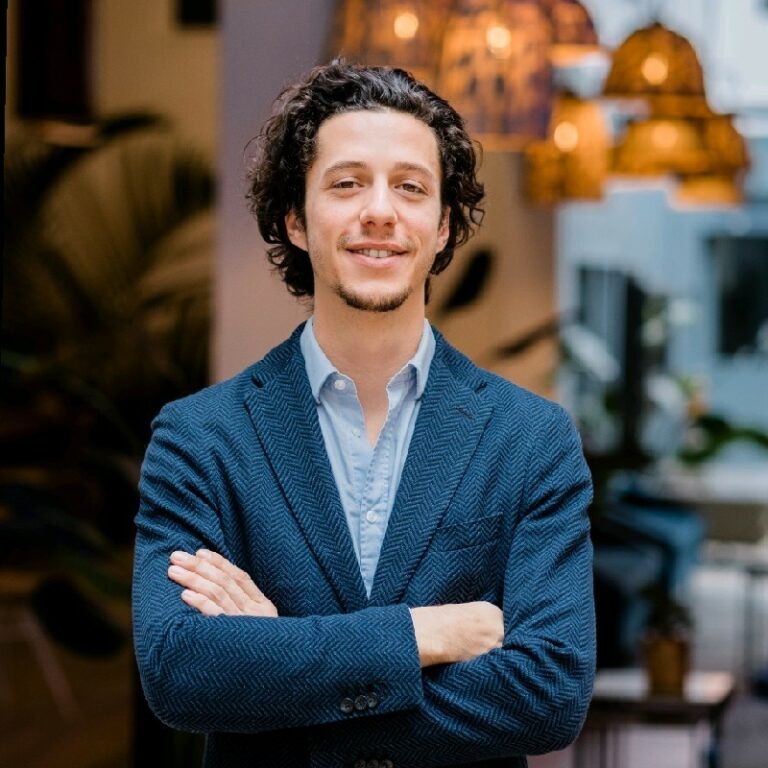 Confident young woman with long blonde hair wearing a tan blazer and white blouse in a modern office setting.
