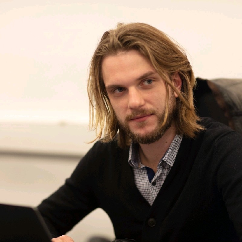 Confident young professional woman with blonde hair wearing a beige blazer and white blouse standing indoors near large windows.