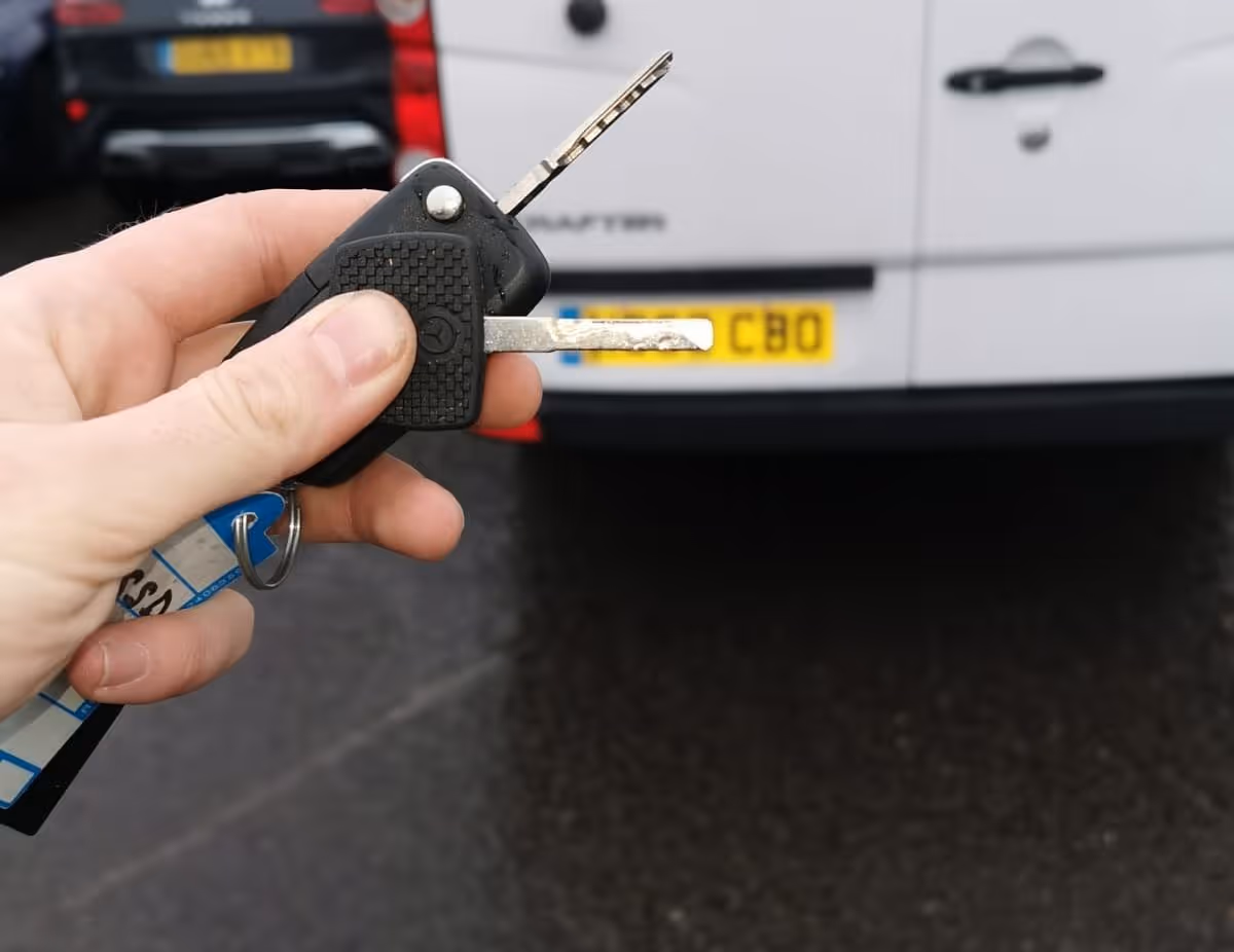 Hand holding a black car key fob with two keys extended in front of a white van with a yellow license plate.