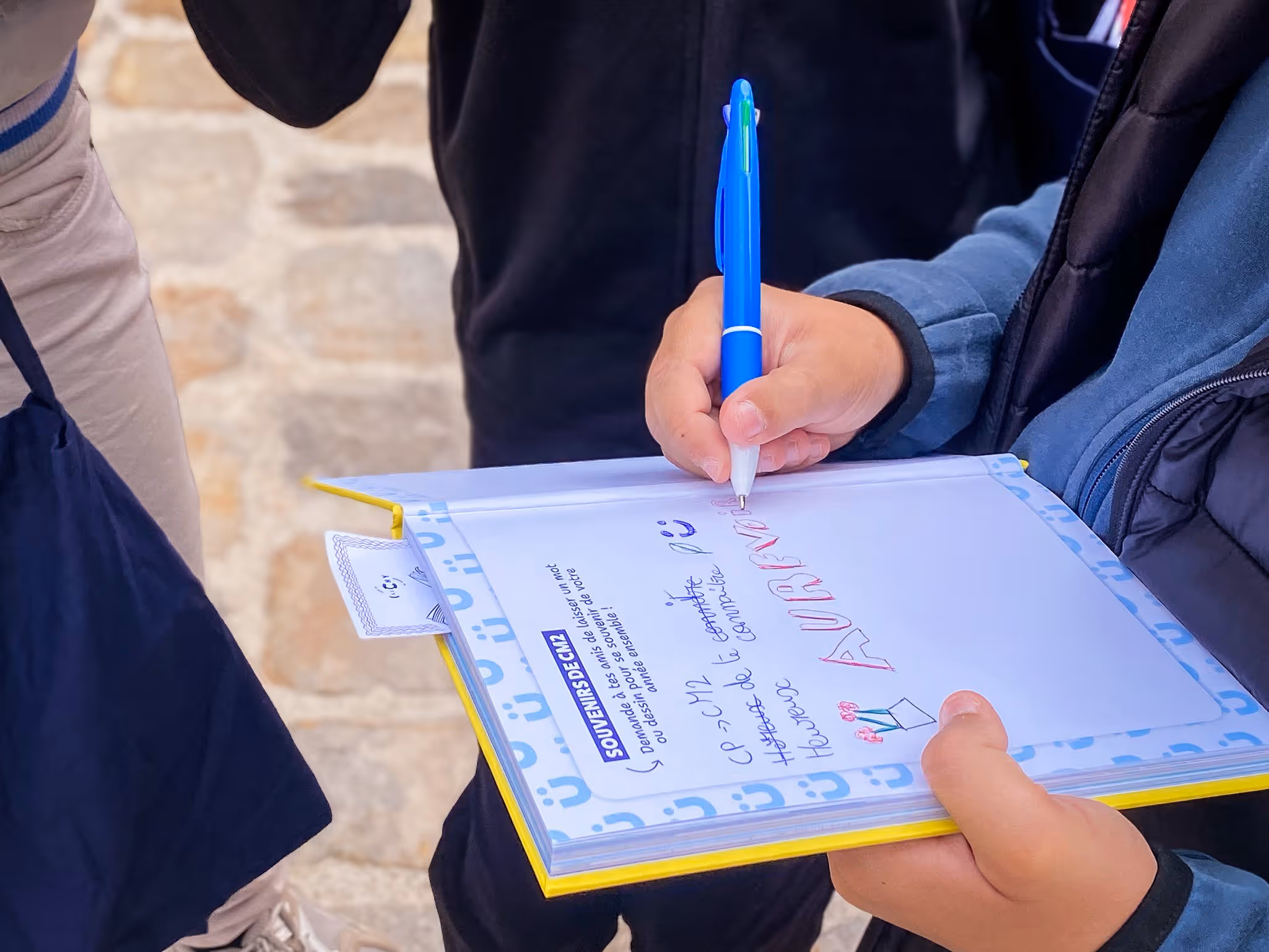 Enfant tenant un stylo bleu et écrivant dans un cahier de souvenirs avec une couverture jaune.