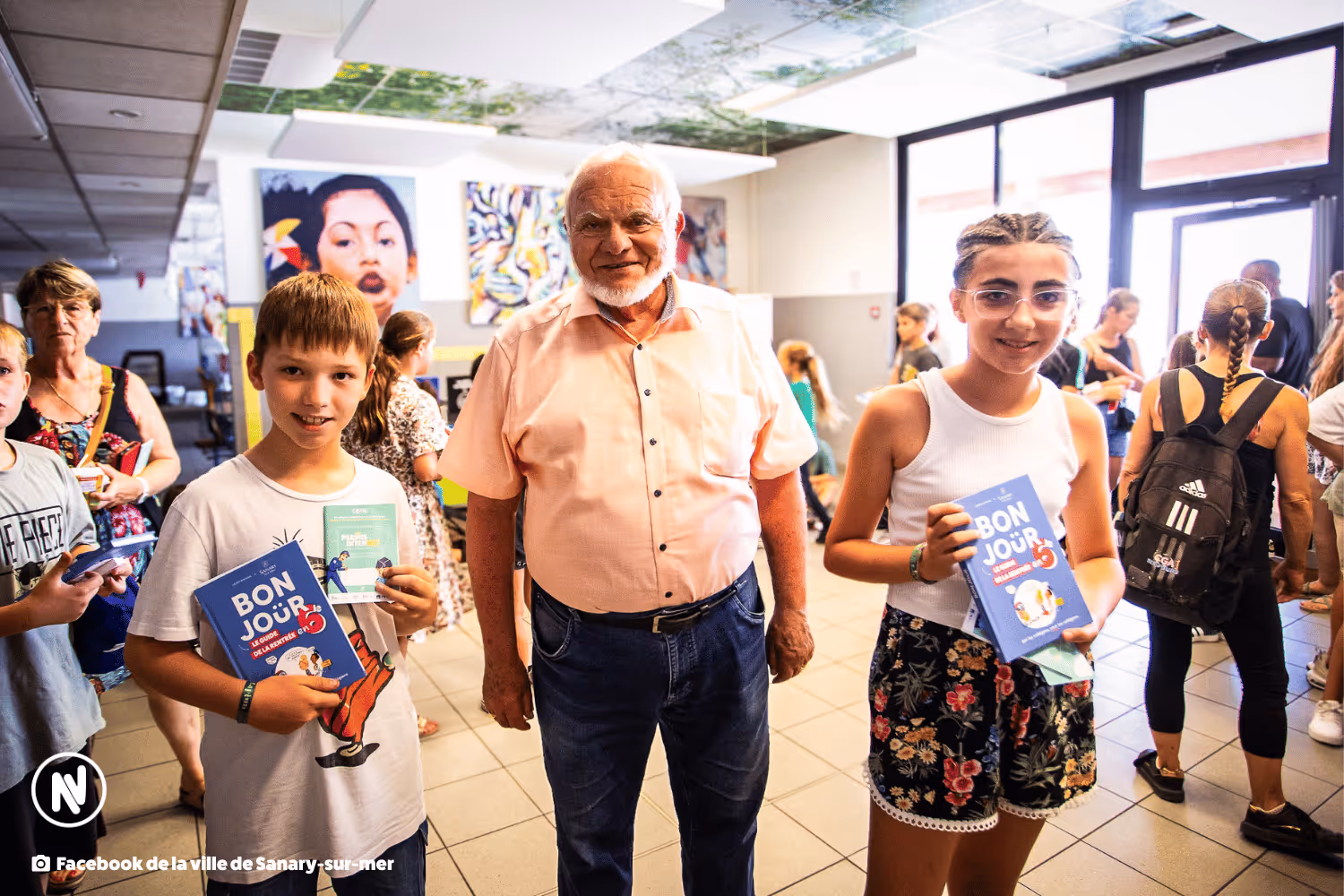 Un garçon et une fille tenant des livres BONJOUR avec un homme âgé souriant entre eux dans un espace intérieur animé.