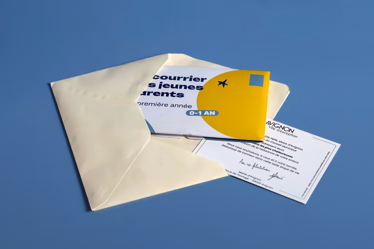 Une enveloppe beige ouverte contenant une carte jaune et blanche avec le texte « Le courrier des jeunes parents 0-1 an » sur fond bleu.