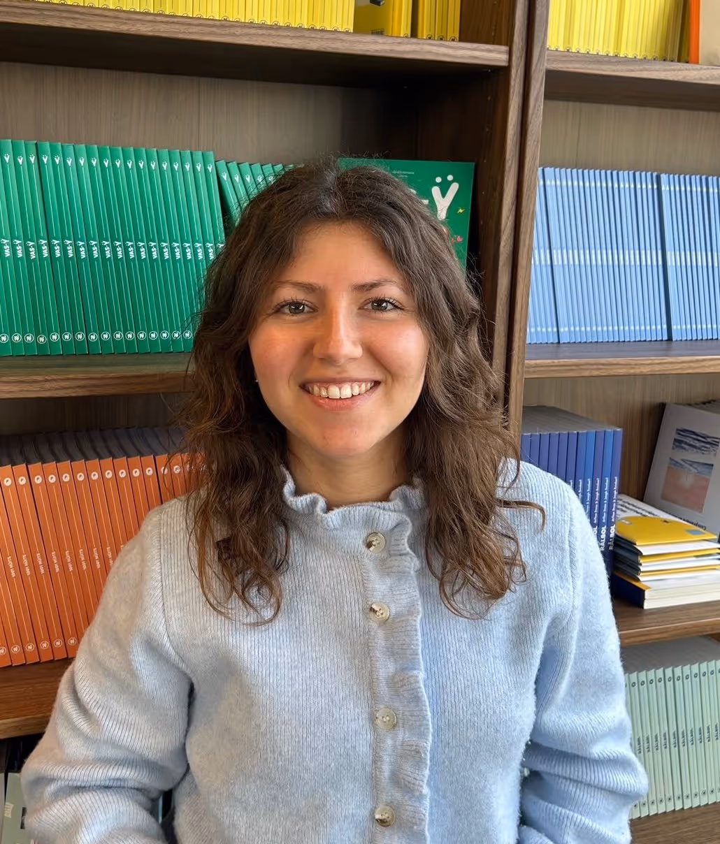 Jeune femme souriante portant un pull gris clair devant une étagère remplie de livres colorés.