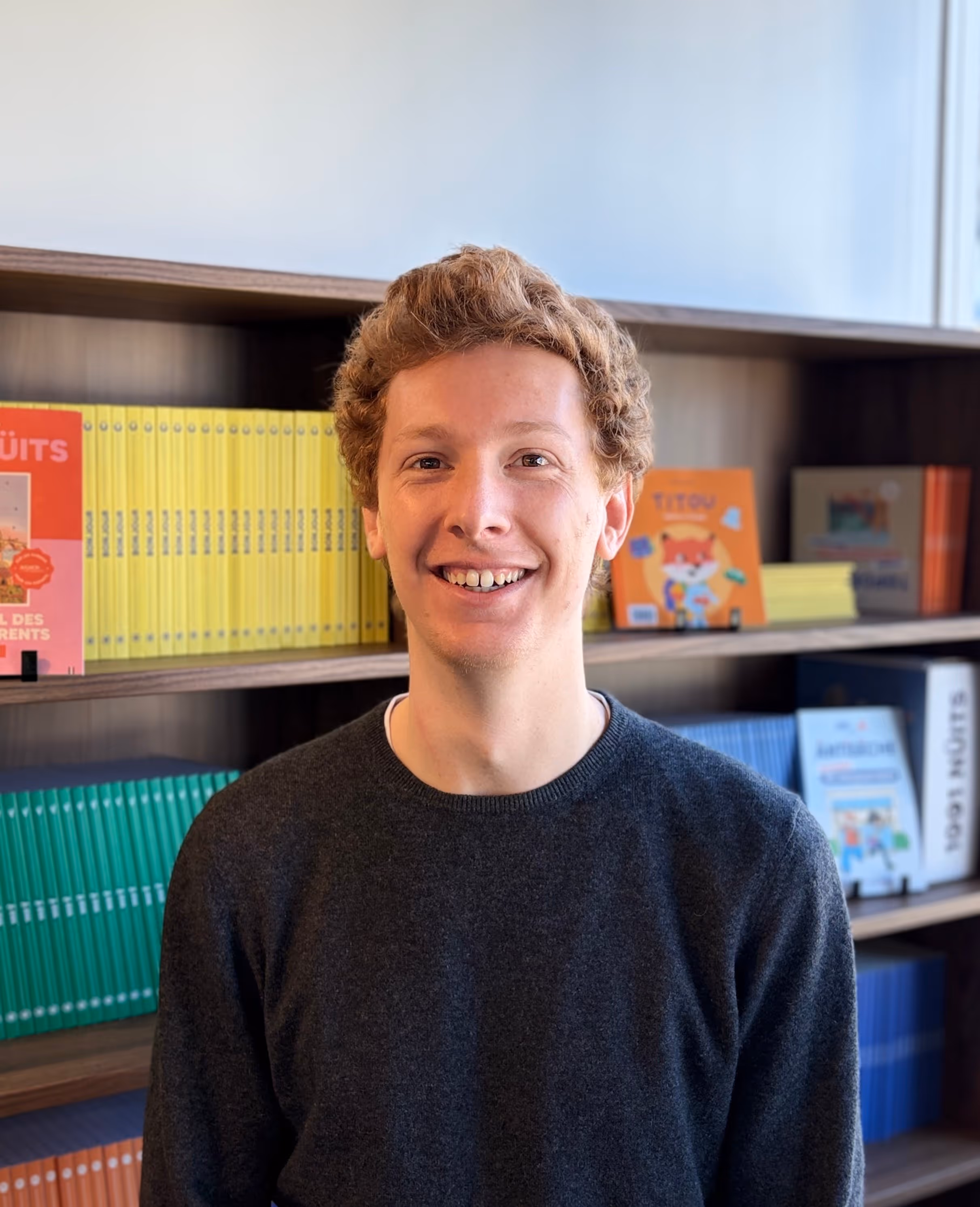 Jeune homme souriant aux cheveux bouclés et pull gris devant une bibliothèque avec des livres colorés.