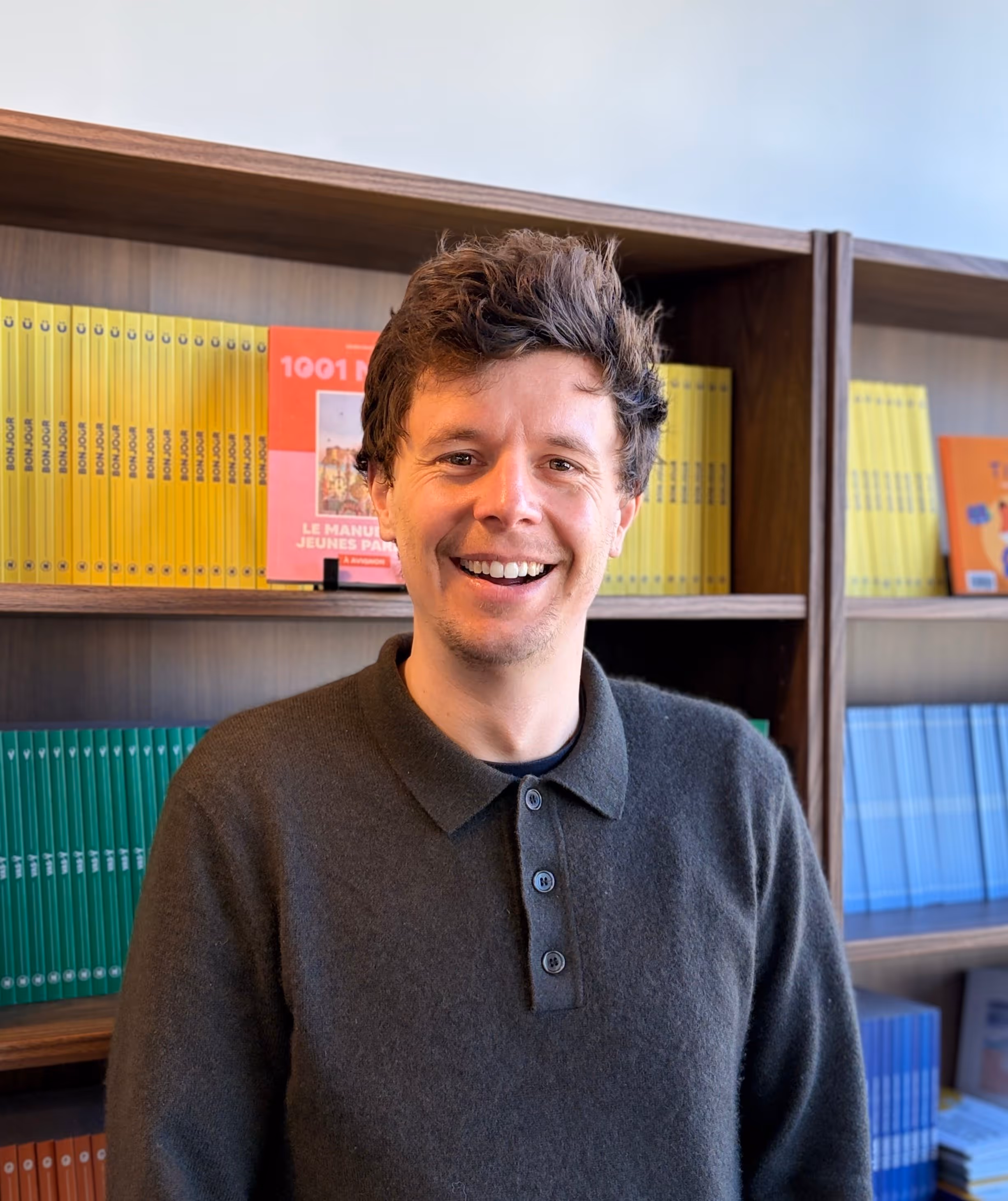 Homme souriant avec un pull gris devant une étagère remplie de livres colorés.