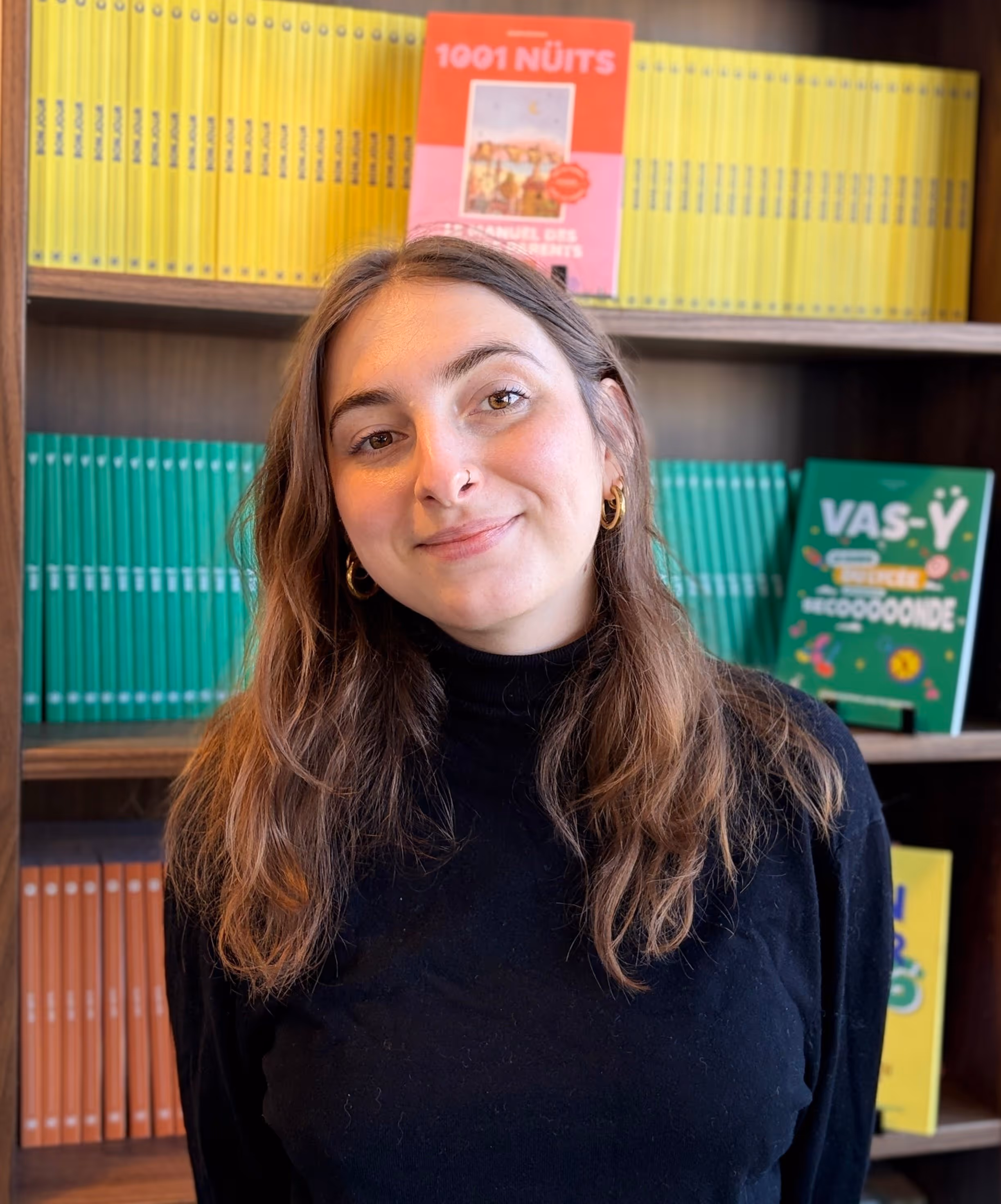 Jeune femme souriante aux cheveux longs portant un pull noir devant une étagère de livres colorés.