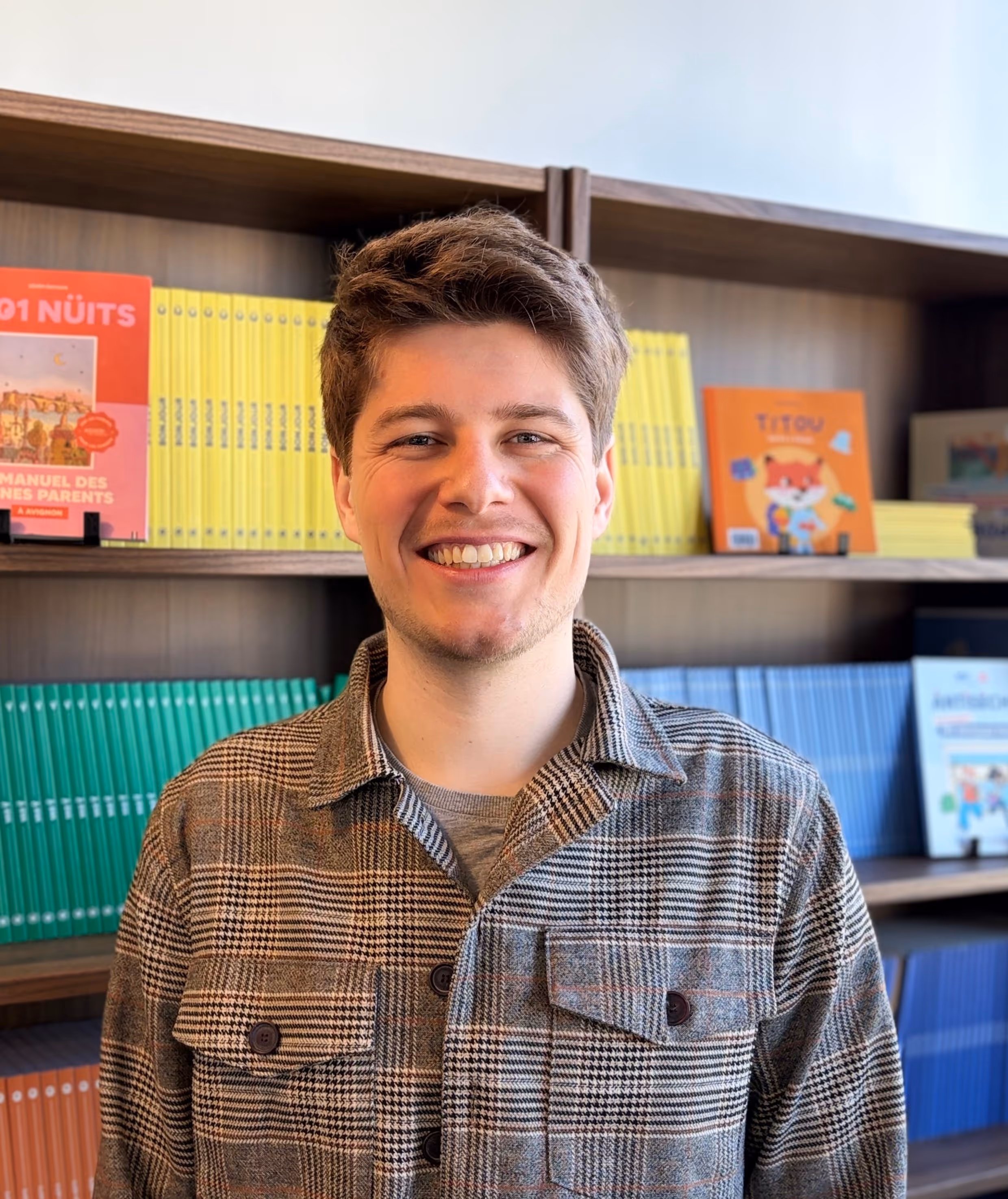 Jeune homme souriant portant une veste à carreaux devant une bibliothèque remplie de livres colorés.