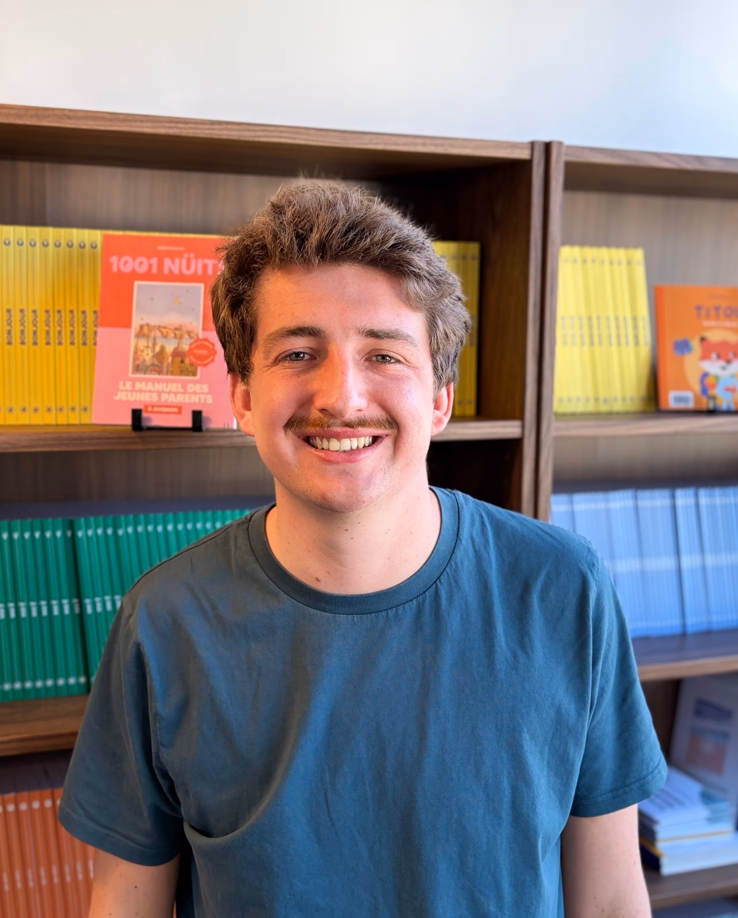 Un homme souriant portant un t-shirt bleu devant une bibliothèque avec des livres colorés.