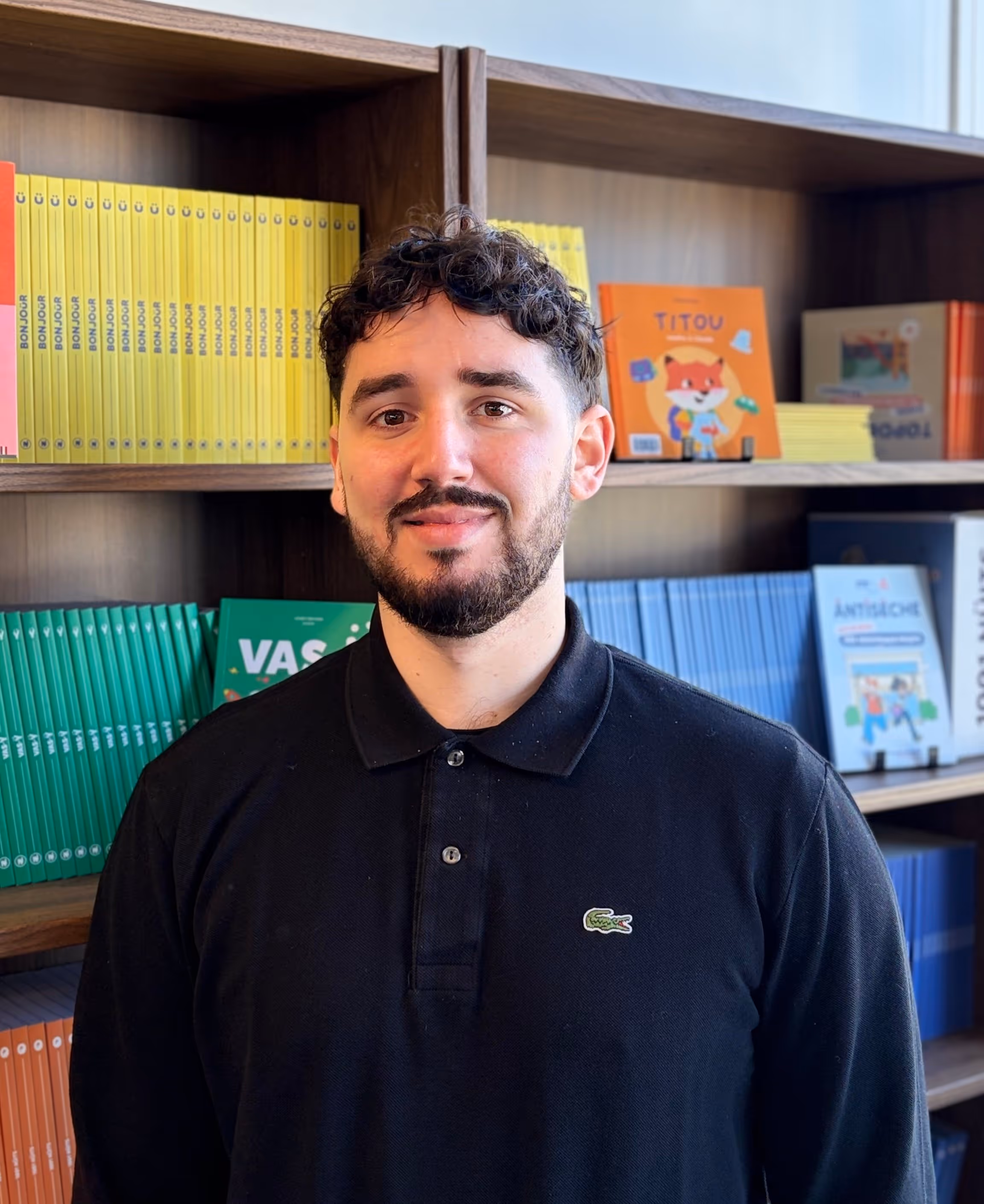 Homme souriant avec une chemise polo noire devant des étagères remplies de livres colorés.