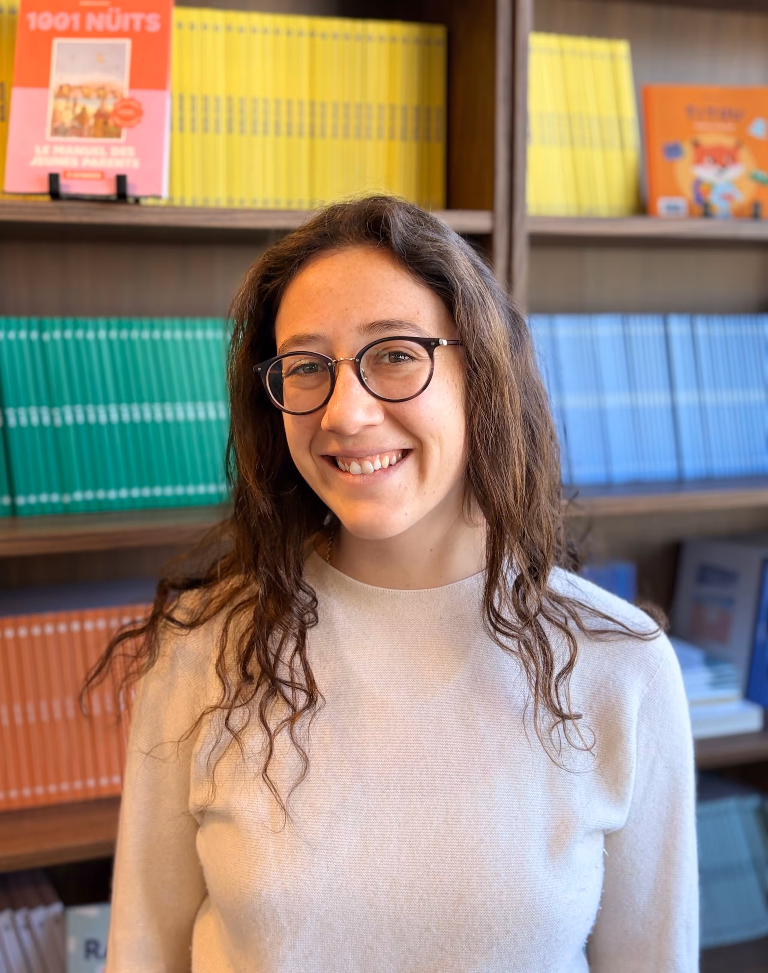 Jeune femme aux cheveux bouclés et lunettes souriant devant une étagère remplie de livres colorés.