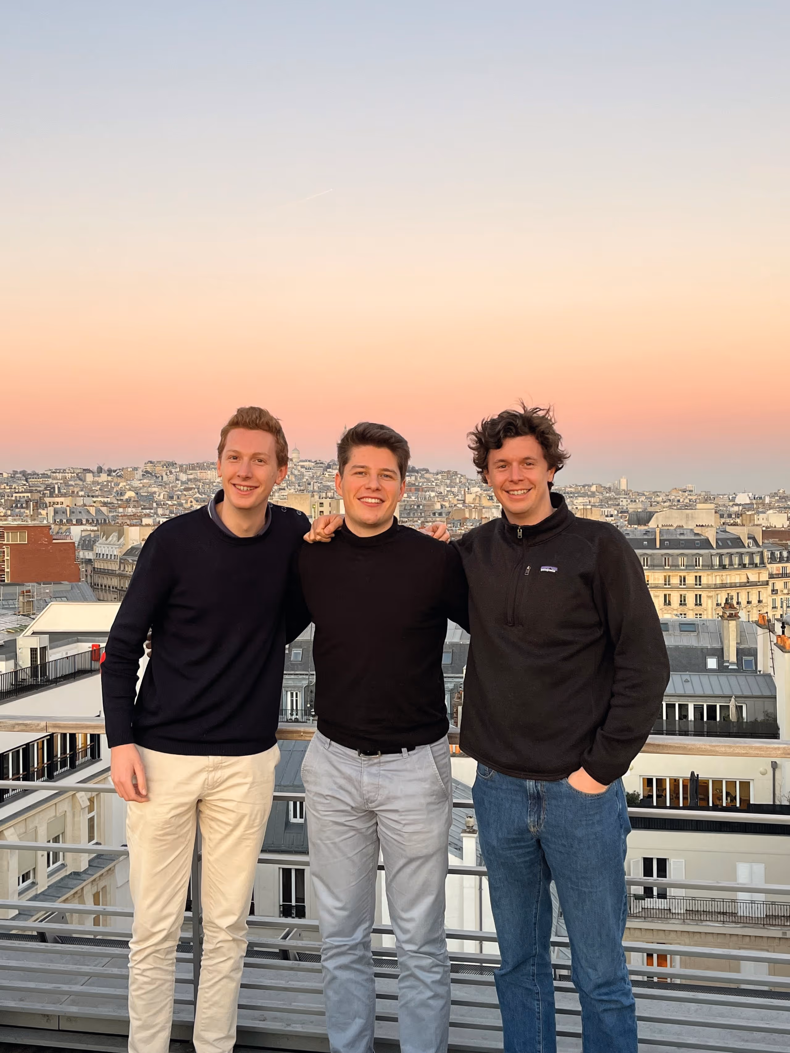 Trois jeunes hommes souriants posant ensemble sur une terrasse avec vue sur les toits de Paris au coucher du soleil.