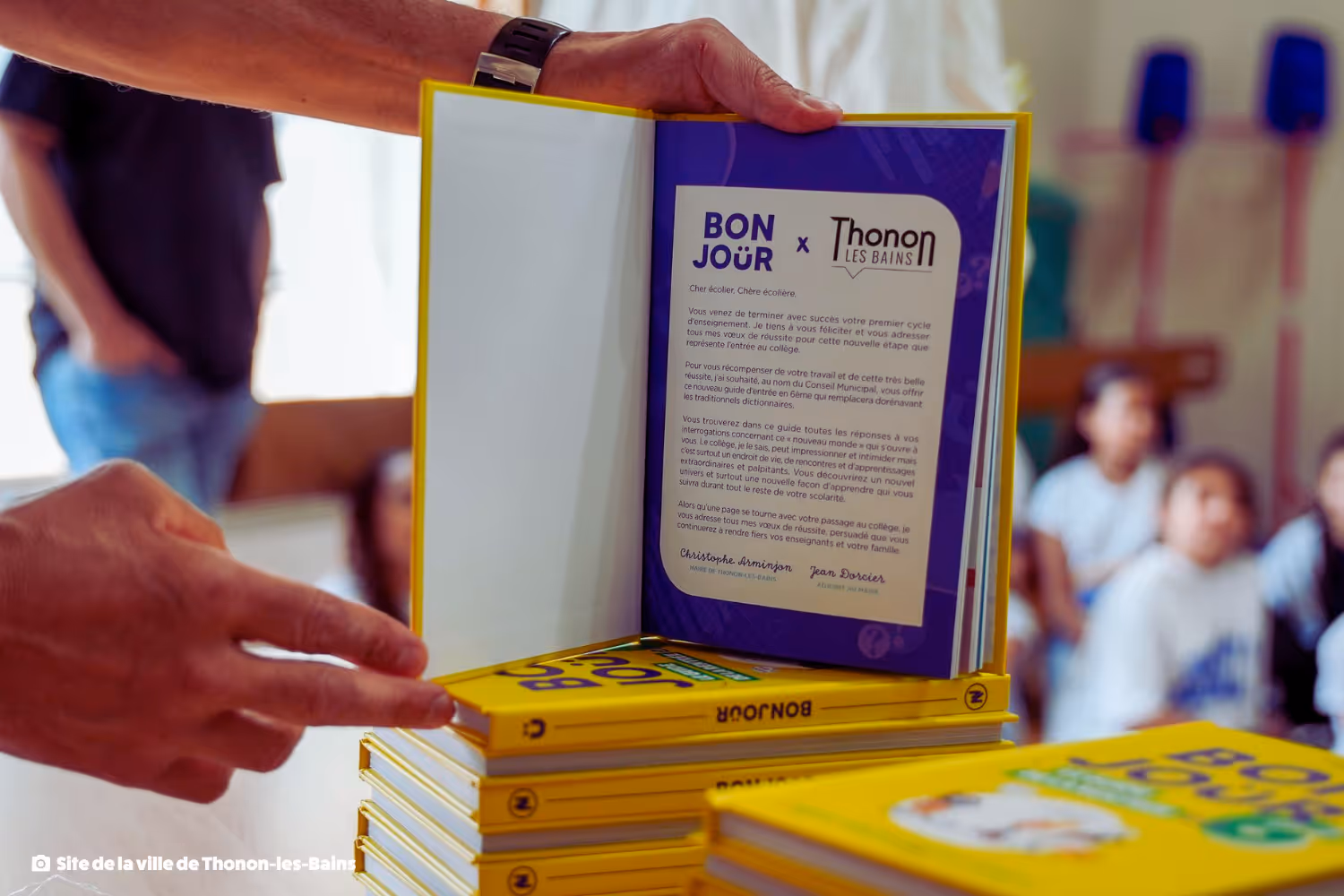 Une personne tient un livre jaune intitulé BONJOUR x Thonon Les Bains ouvert sur une page avec un message imprimé à des élèves en arrière-plan.