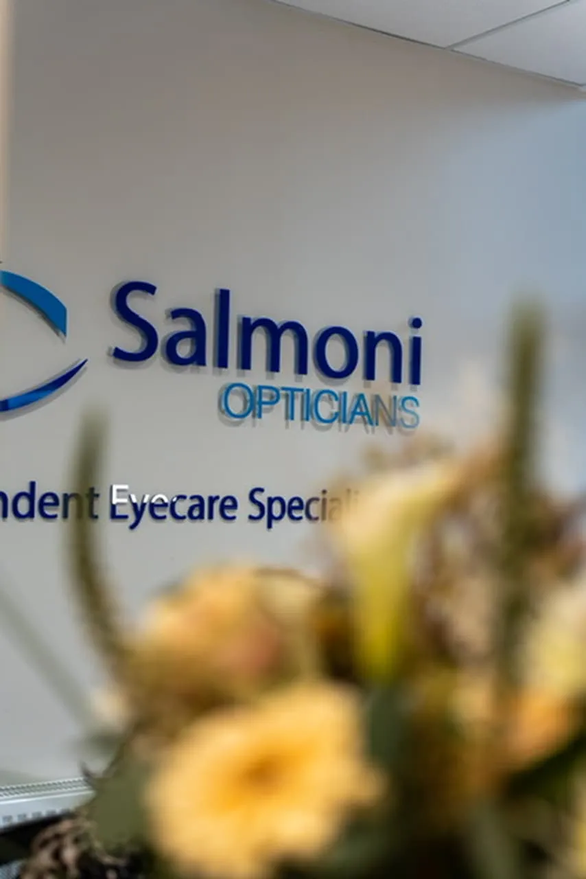 Reception area wall with Salmoni Opticians logo and blurred flowers in foreground.