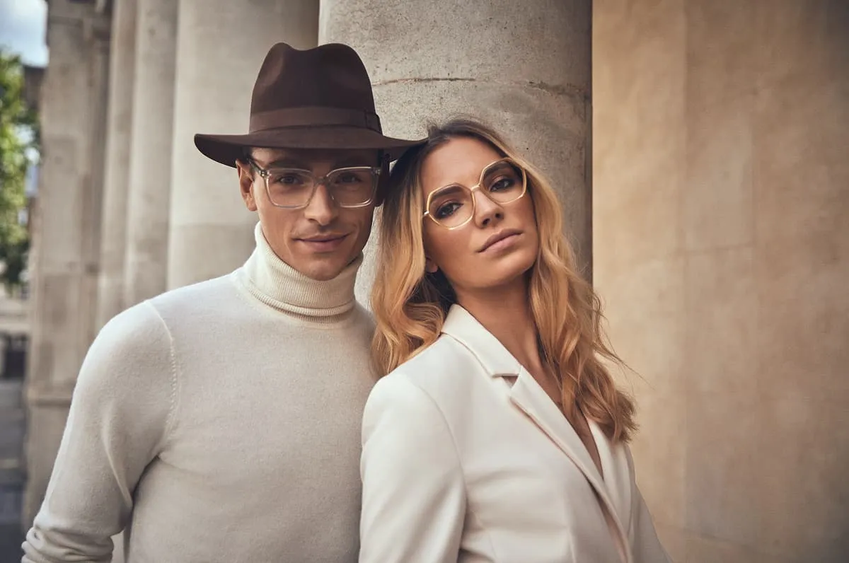 Stylish couple wearing glasses, the man in a brown hat and cream turtleneck, the woman in a white blazer.