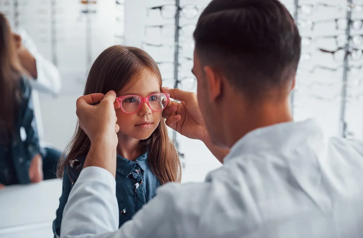 Optician fitting pink glasses on young girl in an eyewear store.