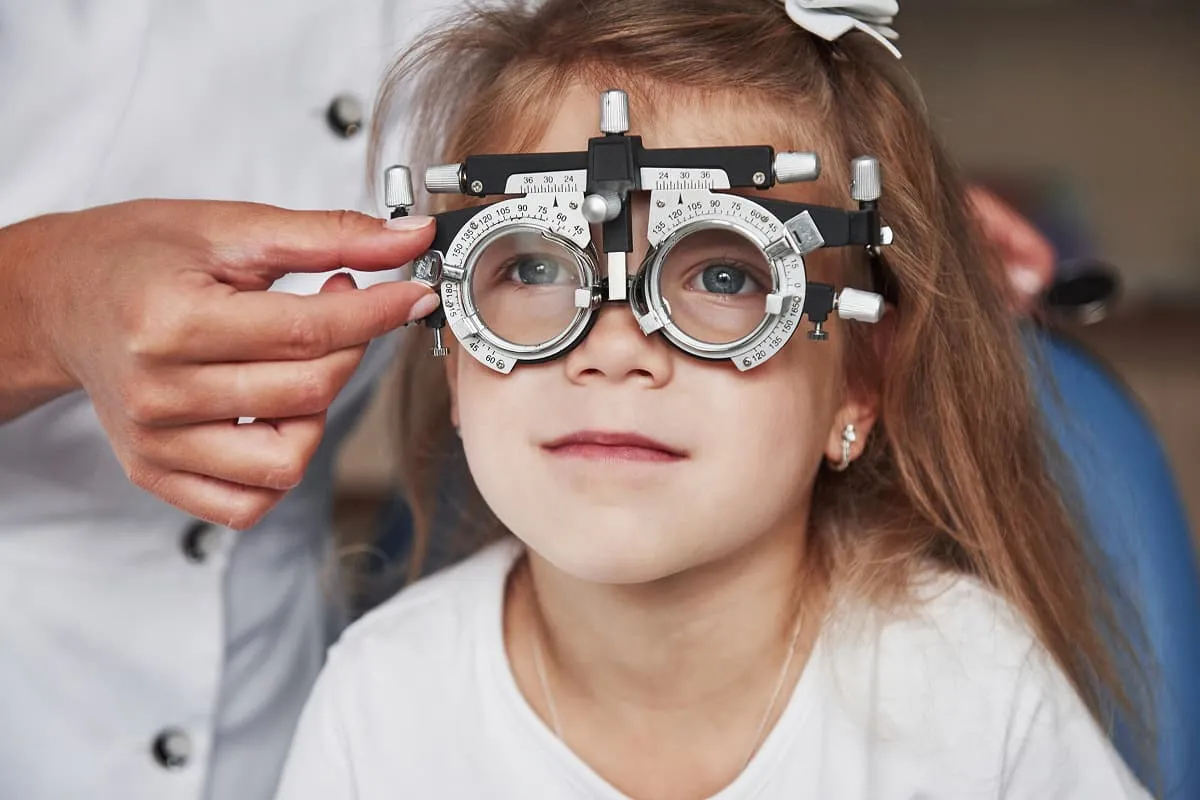 Young girl wearing an eye testing device while an optometrist adjusts it during an eye exam.