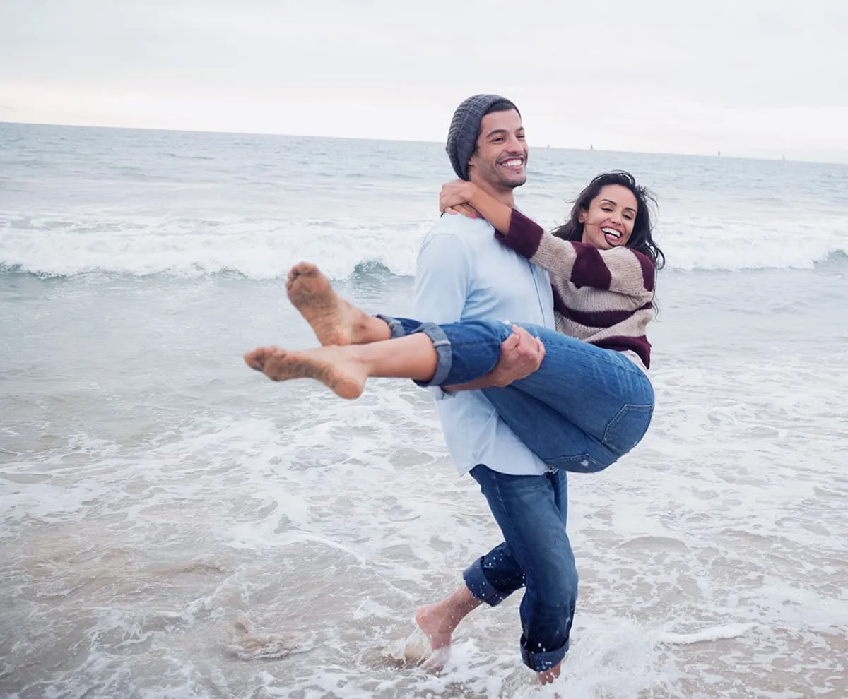 Man wearing jeans and beanie carrying smiling woman in sweater and jeans at the beach with ocean waves.