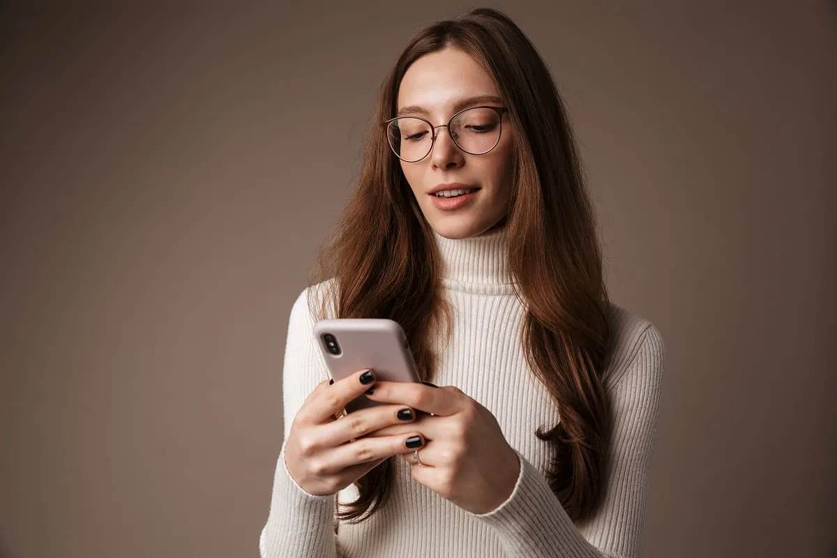 Woman with long brown hair and glasses using a smartphone against a plain brown background.