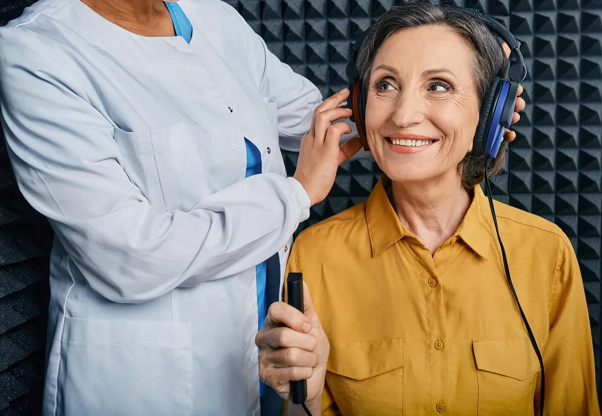 Smiling older woman in a mustard shirt wearing headphones during a hearing test with a healthcare professional.