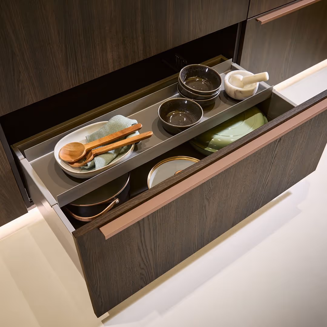 Open kitchen drawer showing stacked plates, bowls, a mortar and pestle, with wooden utensils beside them.