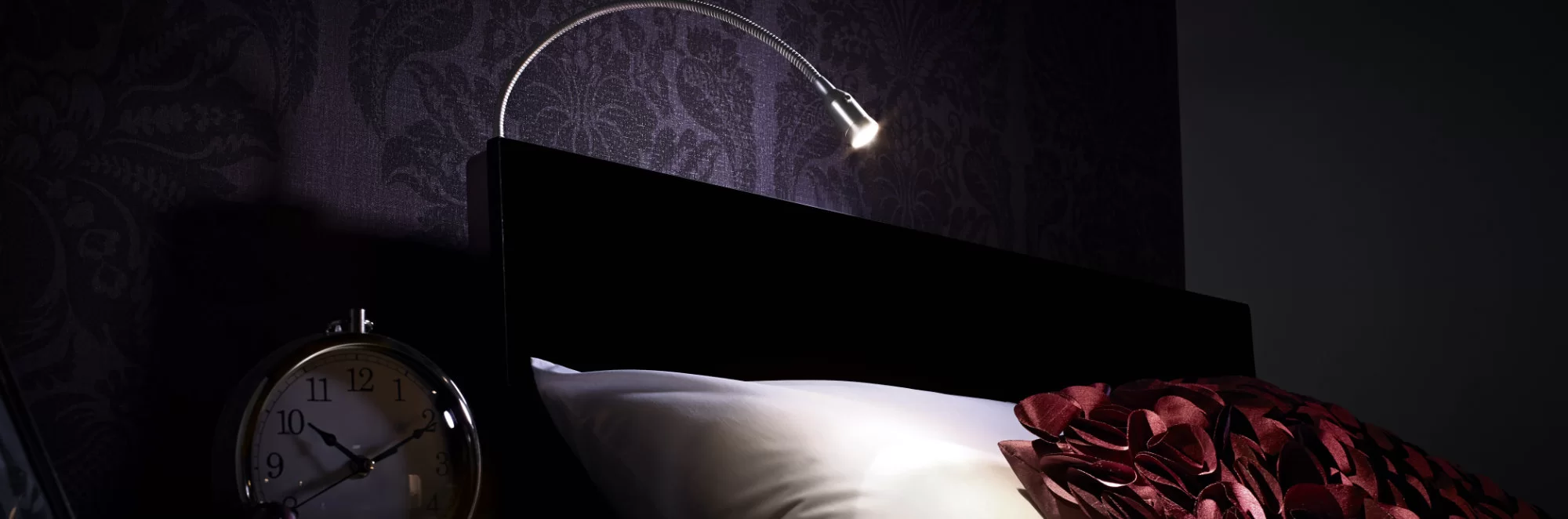 A reading lamp shines on a bed near a clock showing 10:10.