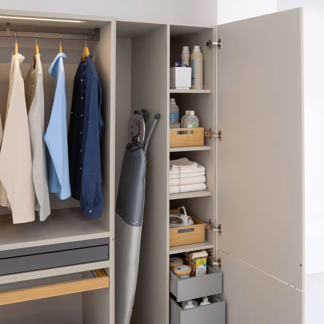An open wardrobe shows shirts, an ironing board, and shelves with cleaning supplies neatly organized.