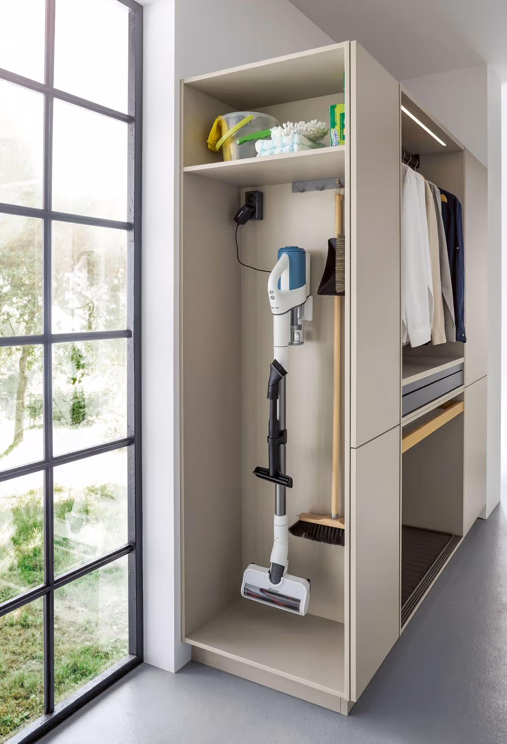 A vacuum cleaner, broom, and cleaning supplies are neatly organized in a tall closet with hanging clothes.