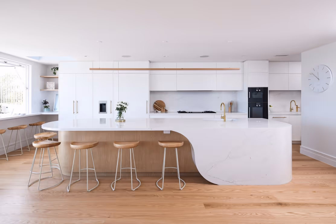Modern kitchen with large white island, wooden bar stools, and sleek cabinets. Bright and clean design.
