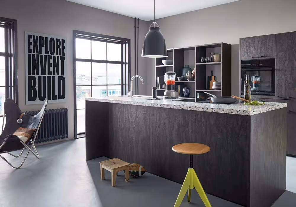 Modern kitchen with a large island, blender, and stool. Wall sign reads "Explore Invent Build."