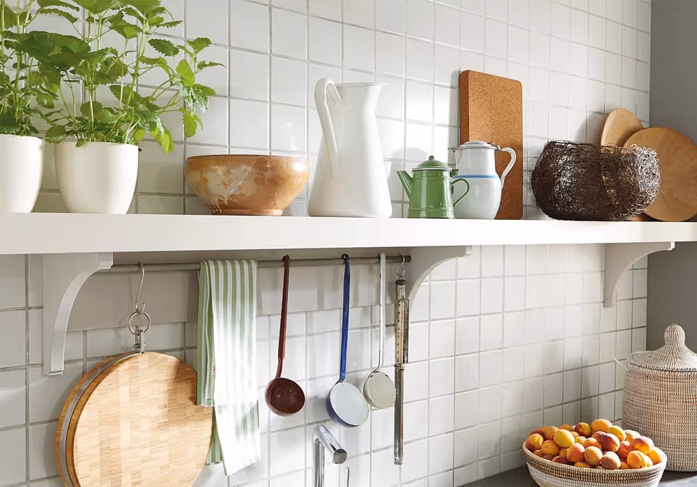Kitchen shelf with plants, bowls, and pitchers. Towels and ladles hang beneath. Baskets hold fruit.