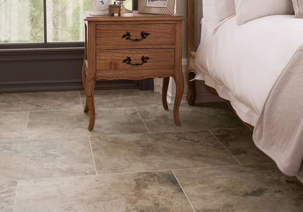 A wooden bedside table with two drawers stands next to a bed on a tiled floor.