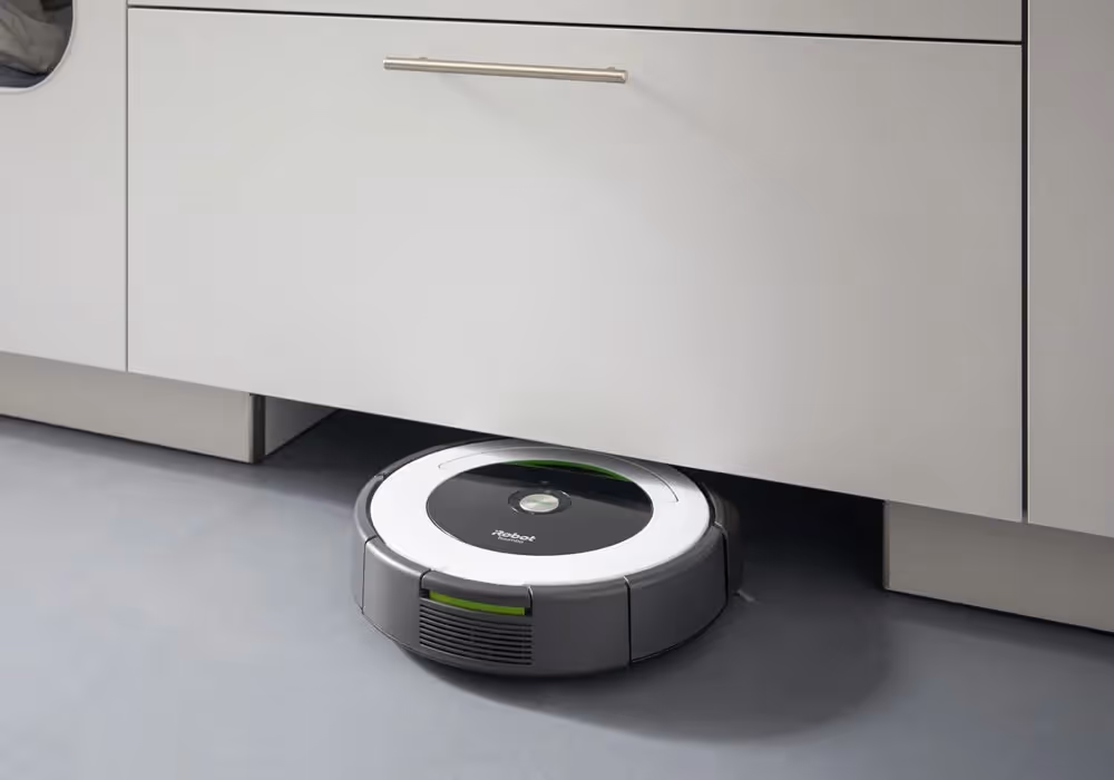A robotic vacuum cleaner moves under a kitchen cabinet on a grey floor.