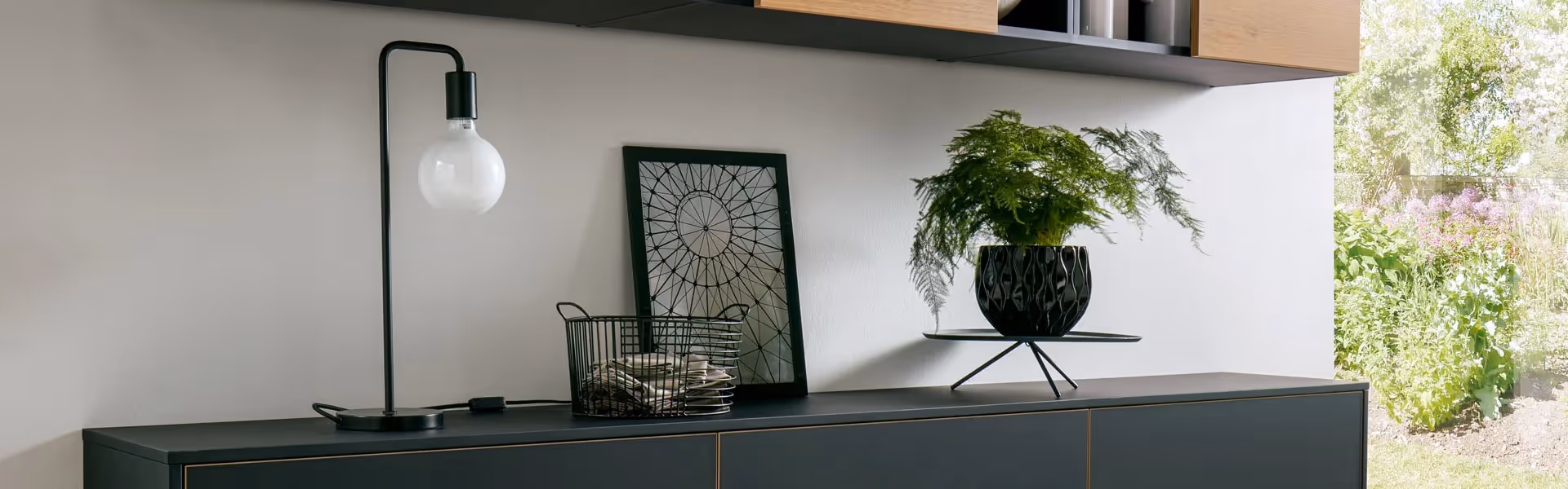 A modern sideboard with a lamp, wire basket, framed artwork, and a potted plant on top.