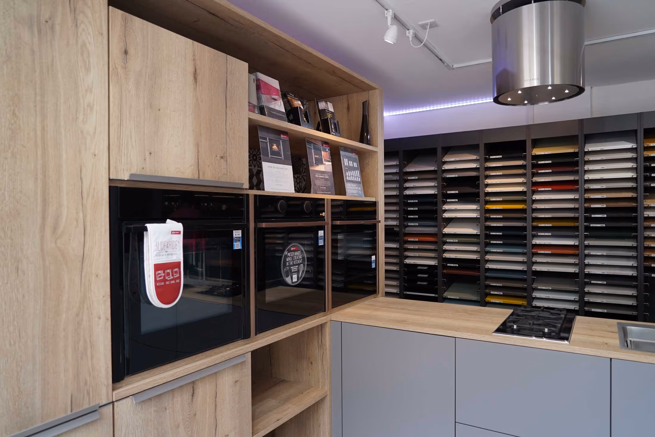 Kitchen showroom displaying modern ovens, stove, and numerous countertop samples in a shelving unit.