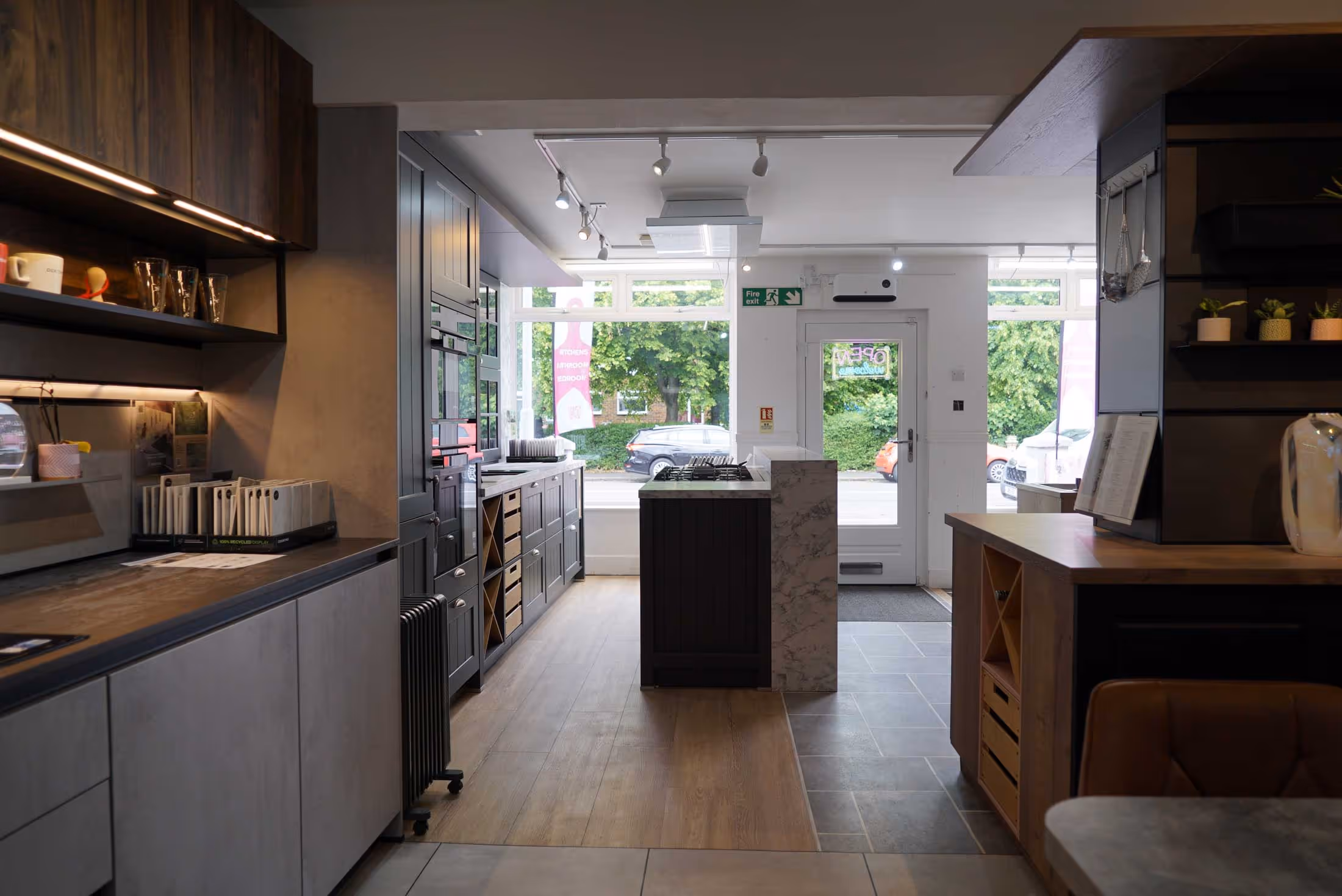A modern kitchen showroom with wooden cabinets, a central island, and bright, natural lighting from large windows.