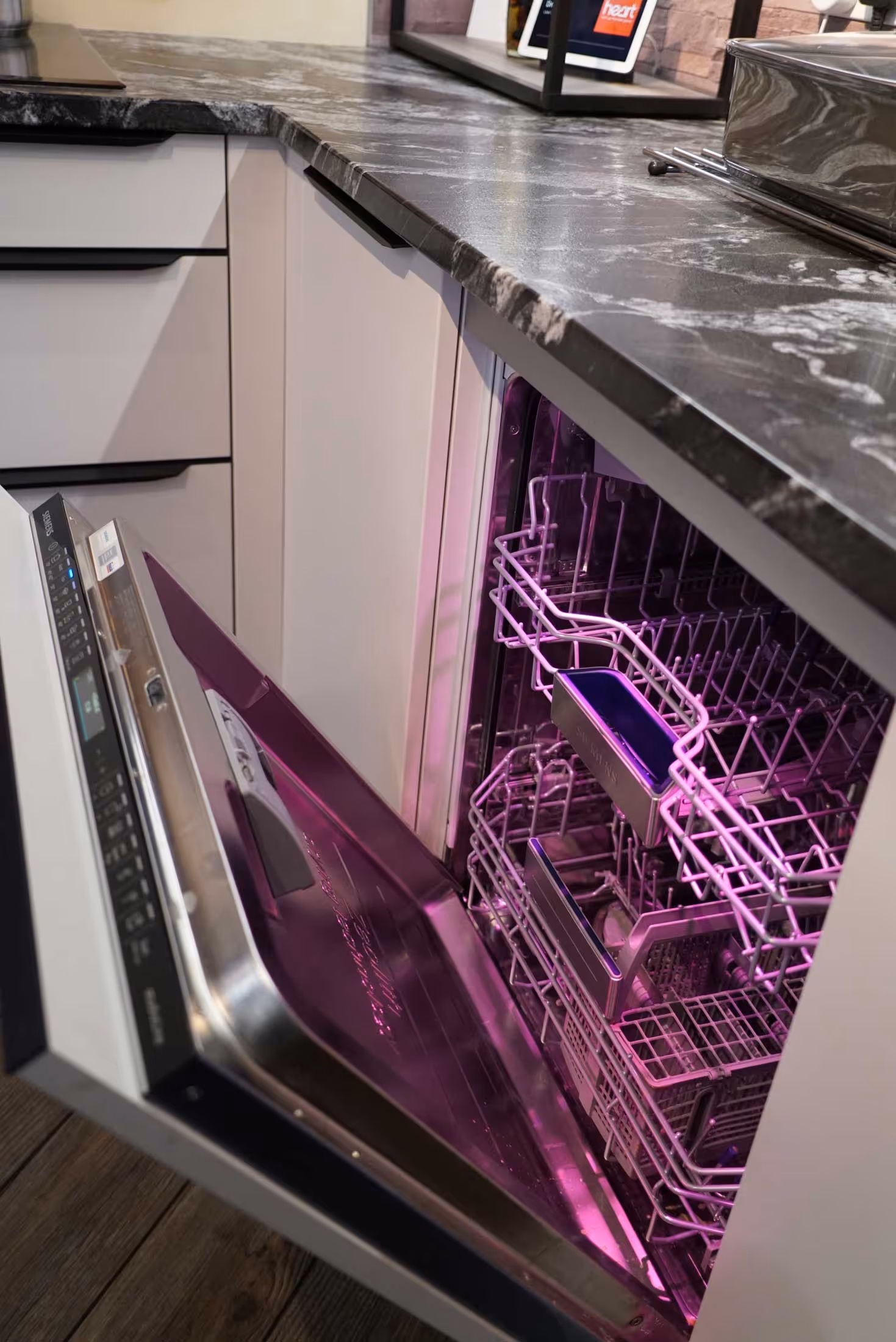 A dishwasher with a pink light is open, revealing empty racks inside.
