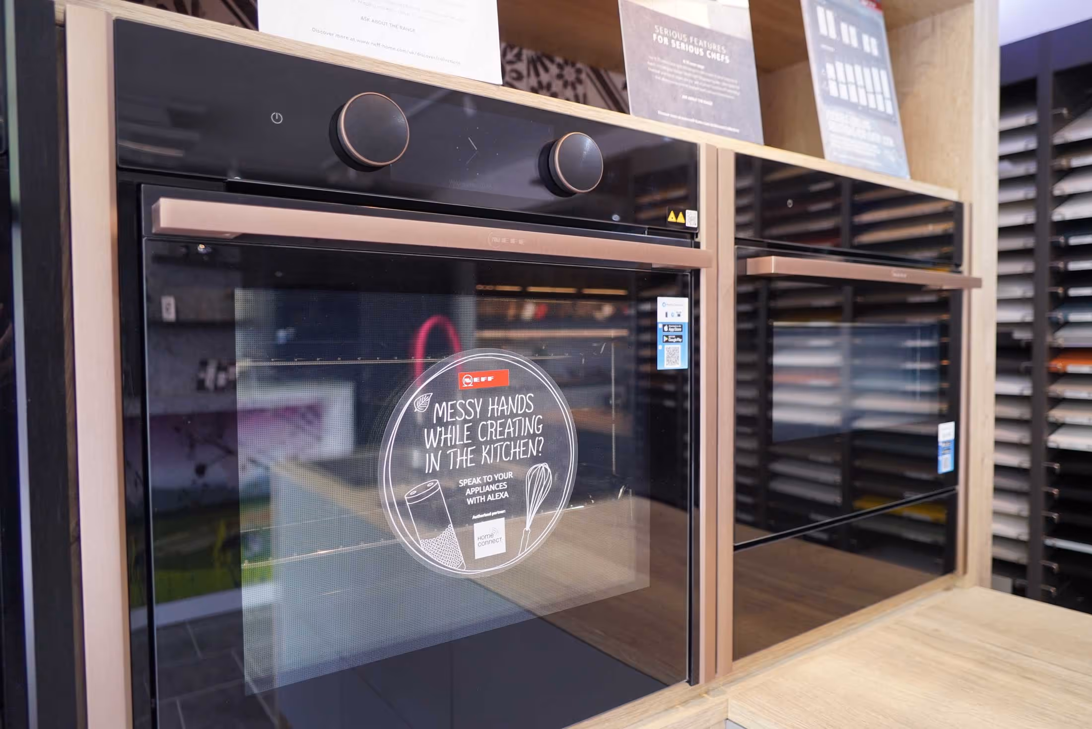 Two modern ovens with digital displays are on a showroom shelf, one has a promotional sticker.