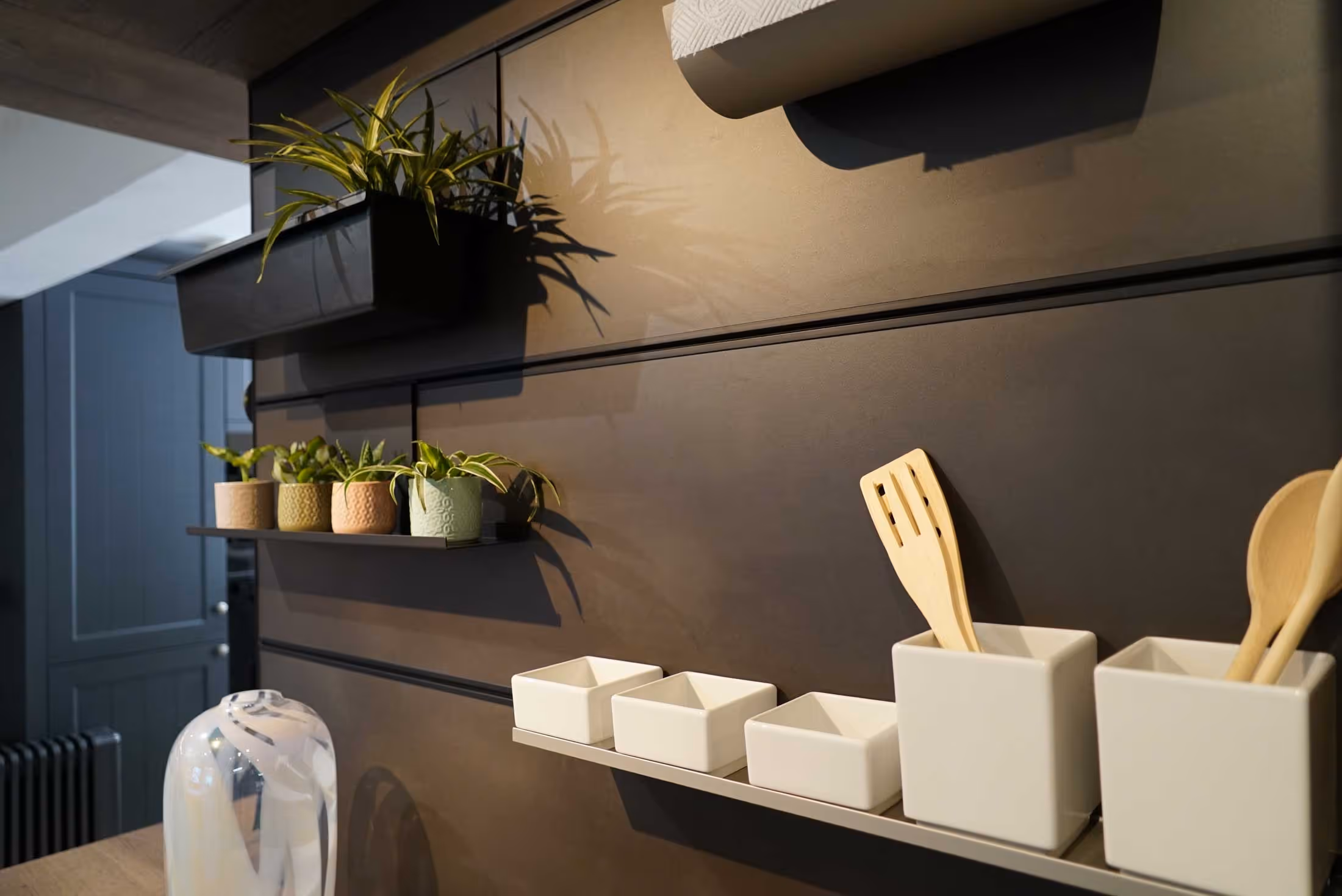 Small plants on shelves and kitchen utensils in containers mounted on a dark wall.