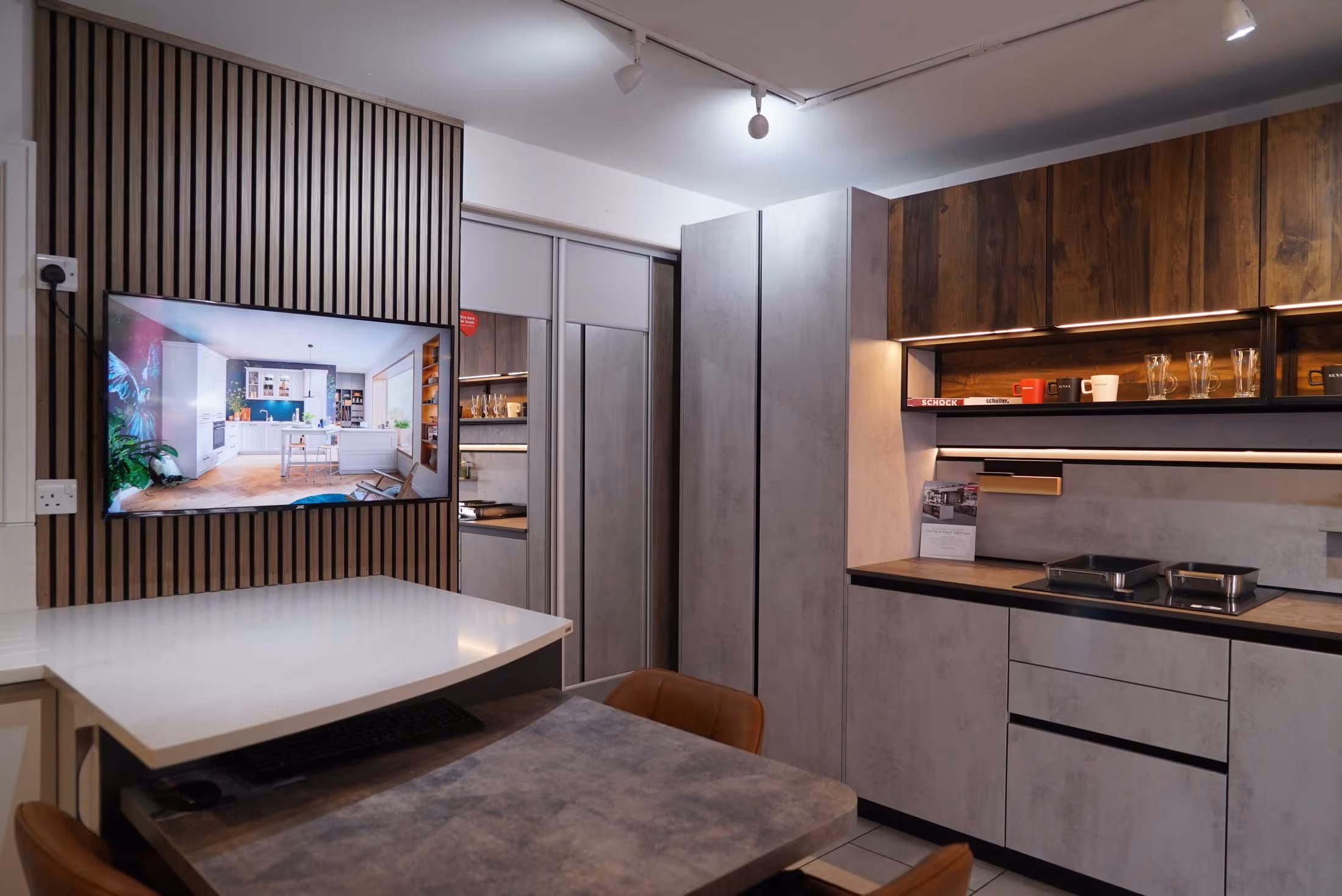Modern kitchen showroom with wooden cabinets, integrated lighting, and a wall-mounted screen displaying a kitchen design.