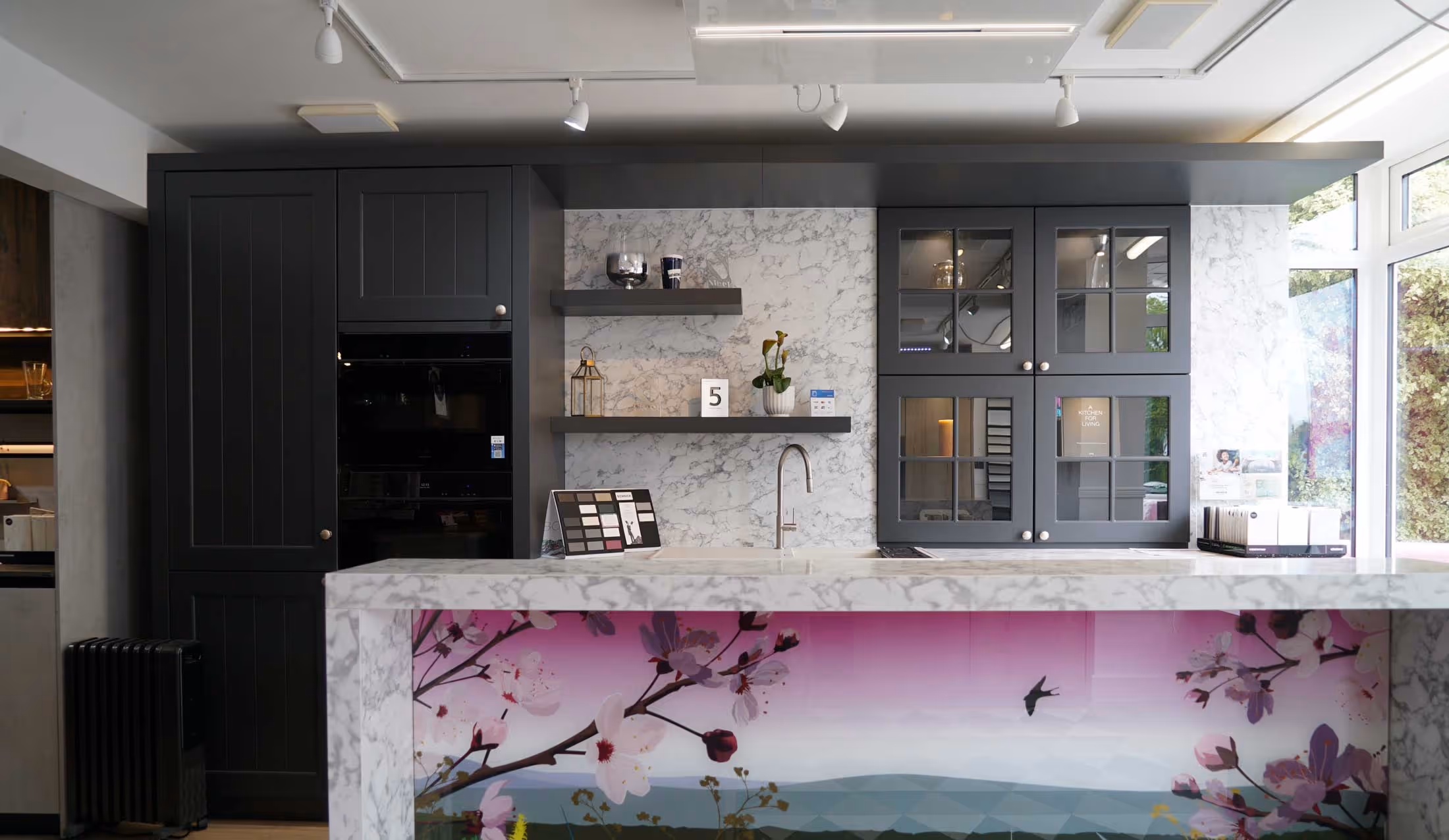 A modern kitchen with dark cabinets, marble countertops, and an island featuring floral cherry blossom artwork.