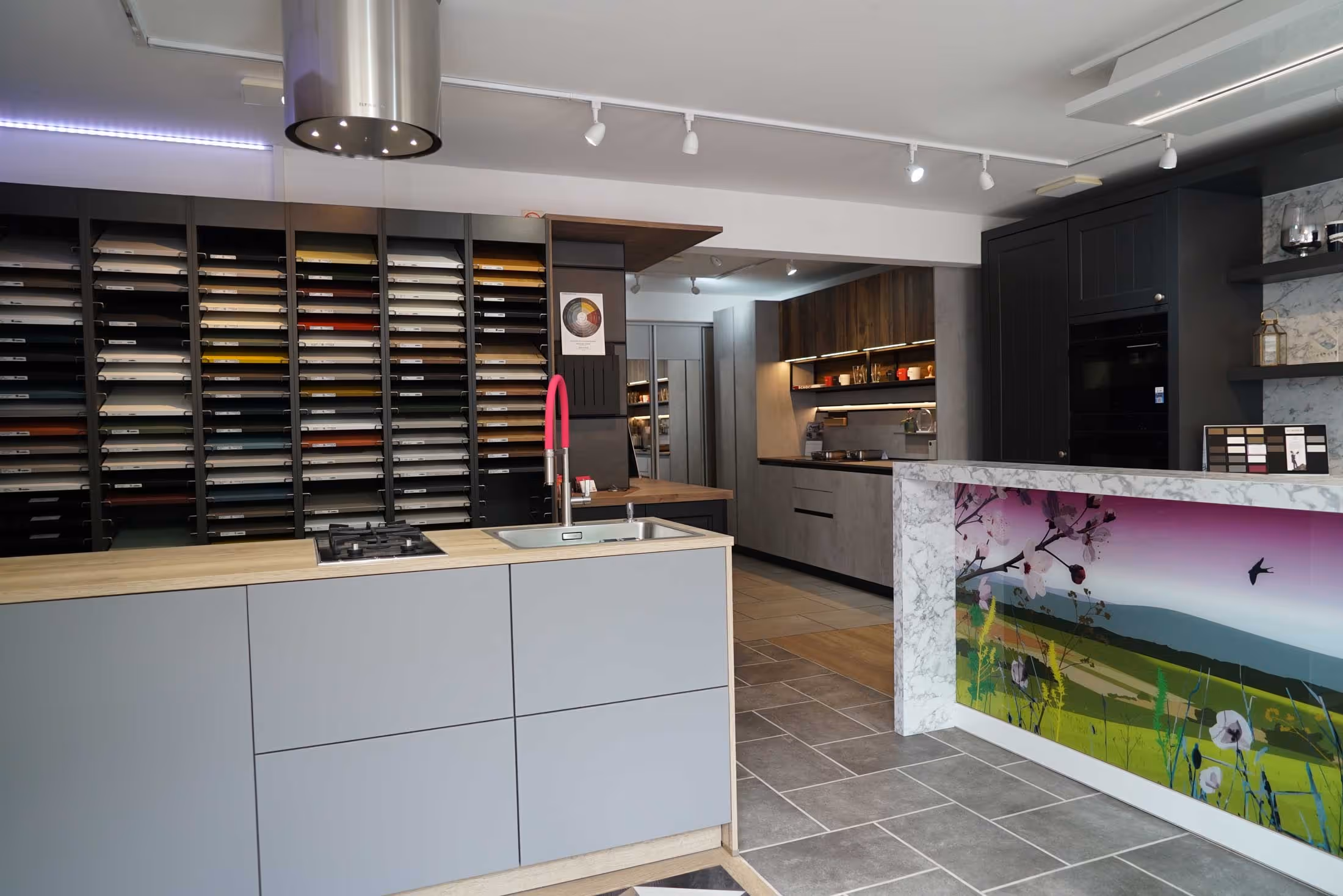 Modern kitchen showroom with various cabinet samples and countertops. Bright light highlights a central island.