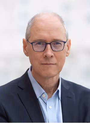 Portrait of a bald man with glasses wearing a dark suit and light blue shirt against a light background.