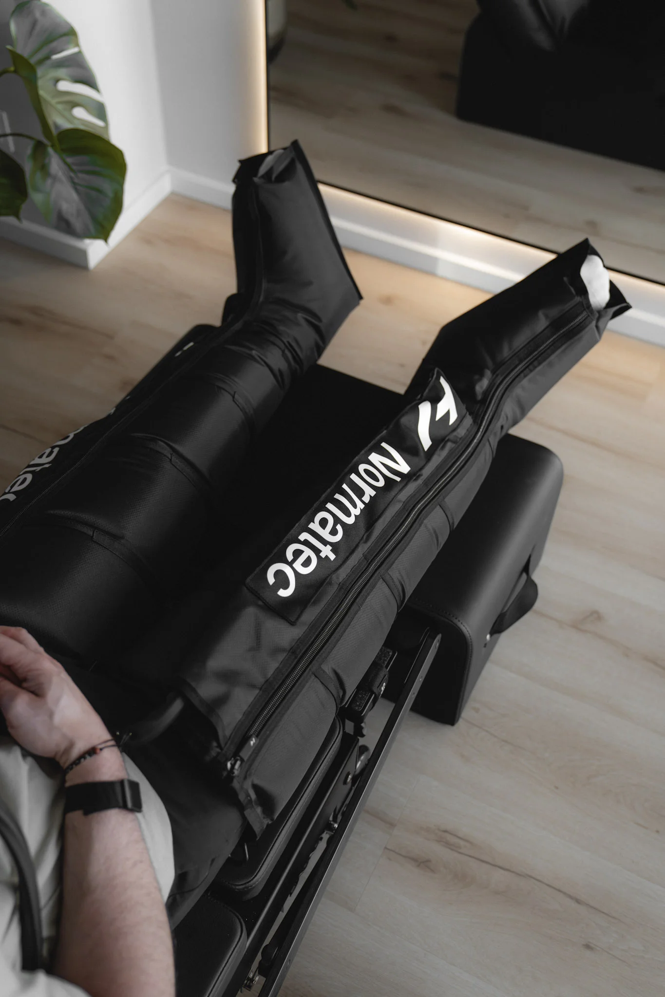 Beine in schwarzen Normatec-Kompressionsstiefeln auf einer schwarzen Liege in einem hellen Raum mit Holzboden.