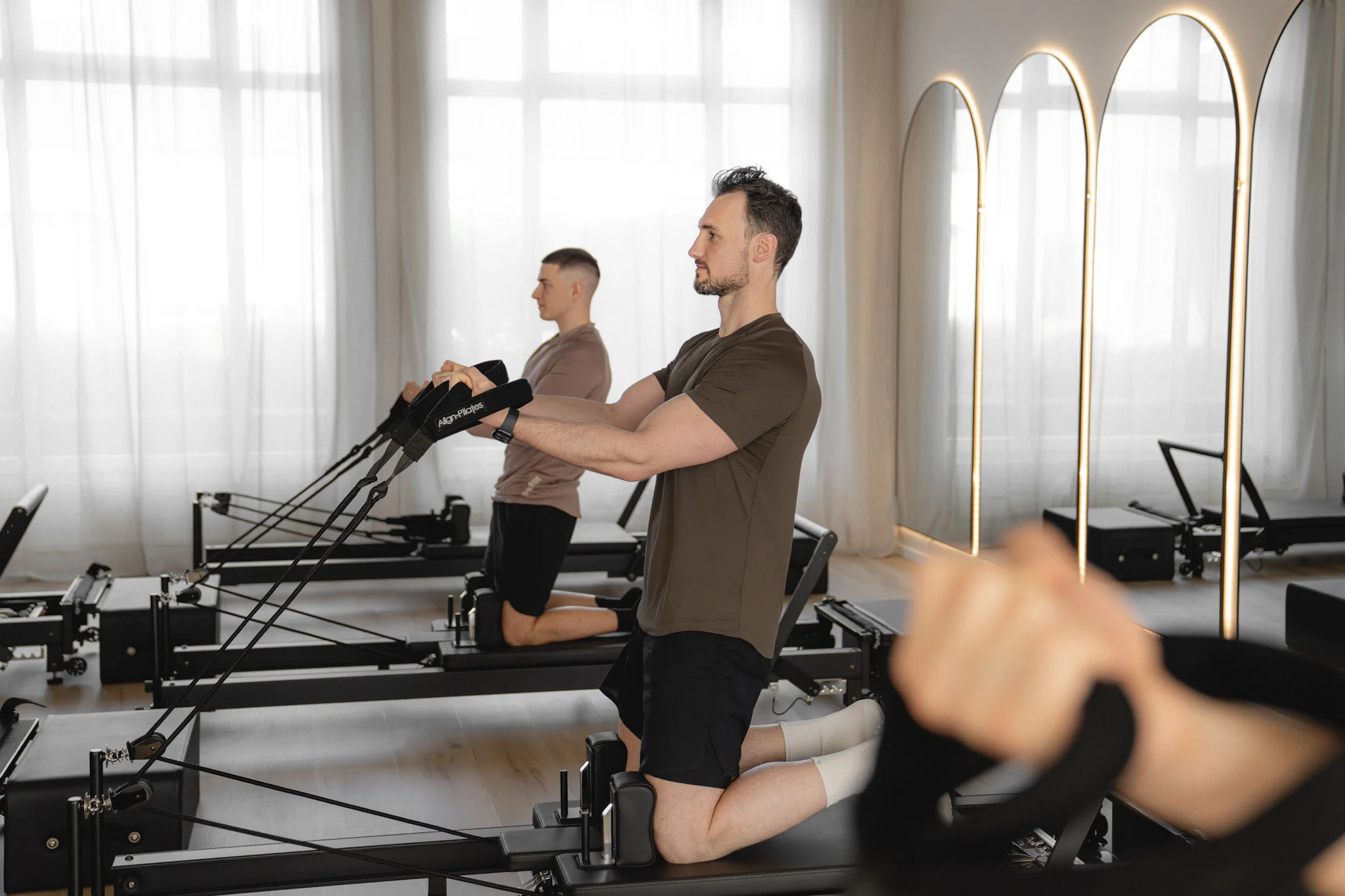 Zwei Männer trainieren auf Pilates-Reformern in einem hellen Pilatesstudio mit großen Fenstern und beleuchteten Spiegeln.