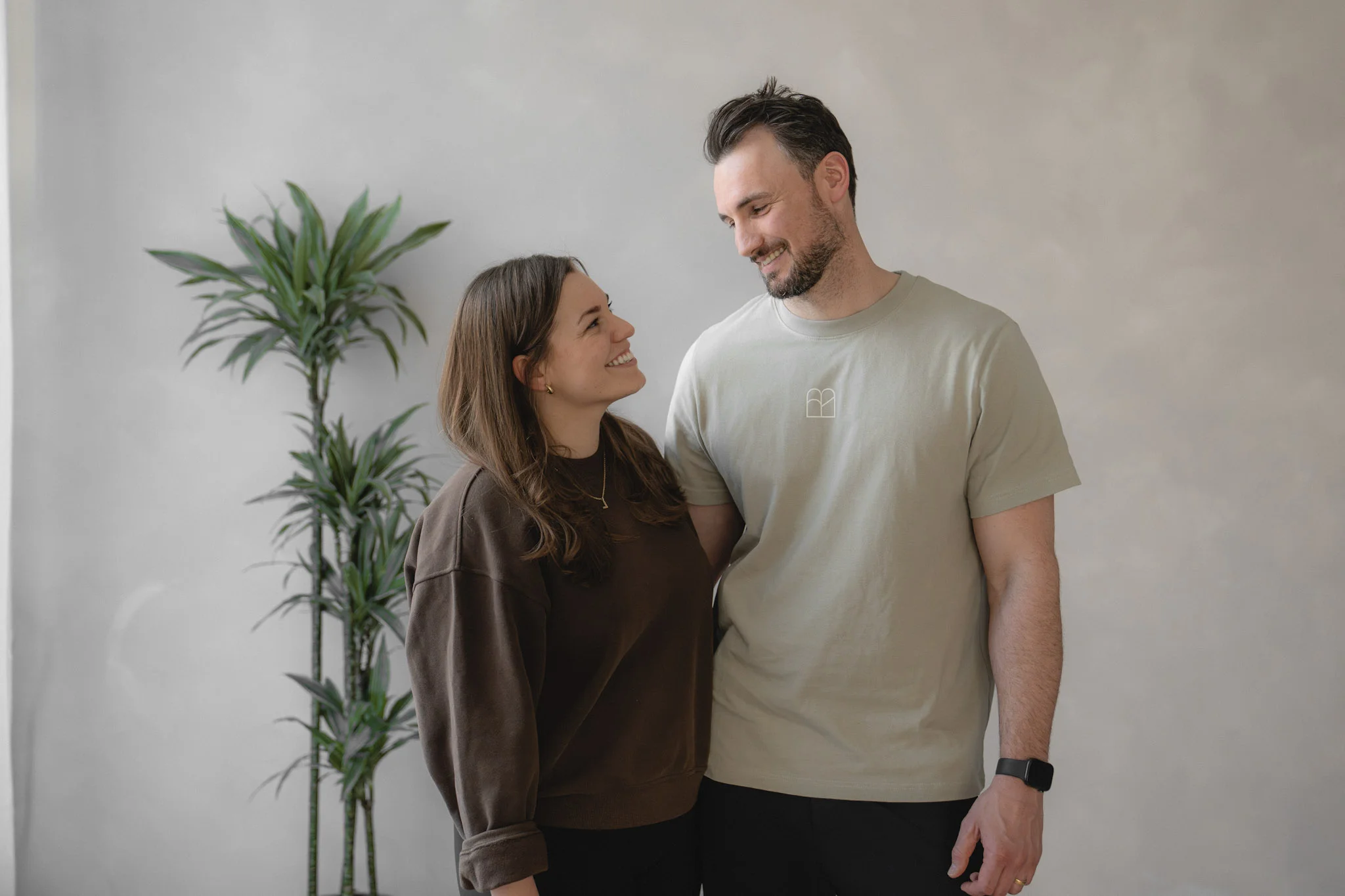 Ein lächelndes Paar blickt sich liebevoll an, stehend vor einer grauen Wand mit einer grünen Pflanze im Hintergrund.