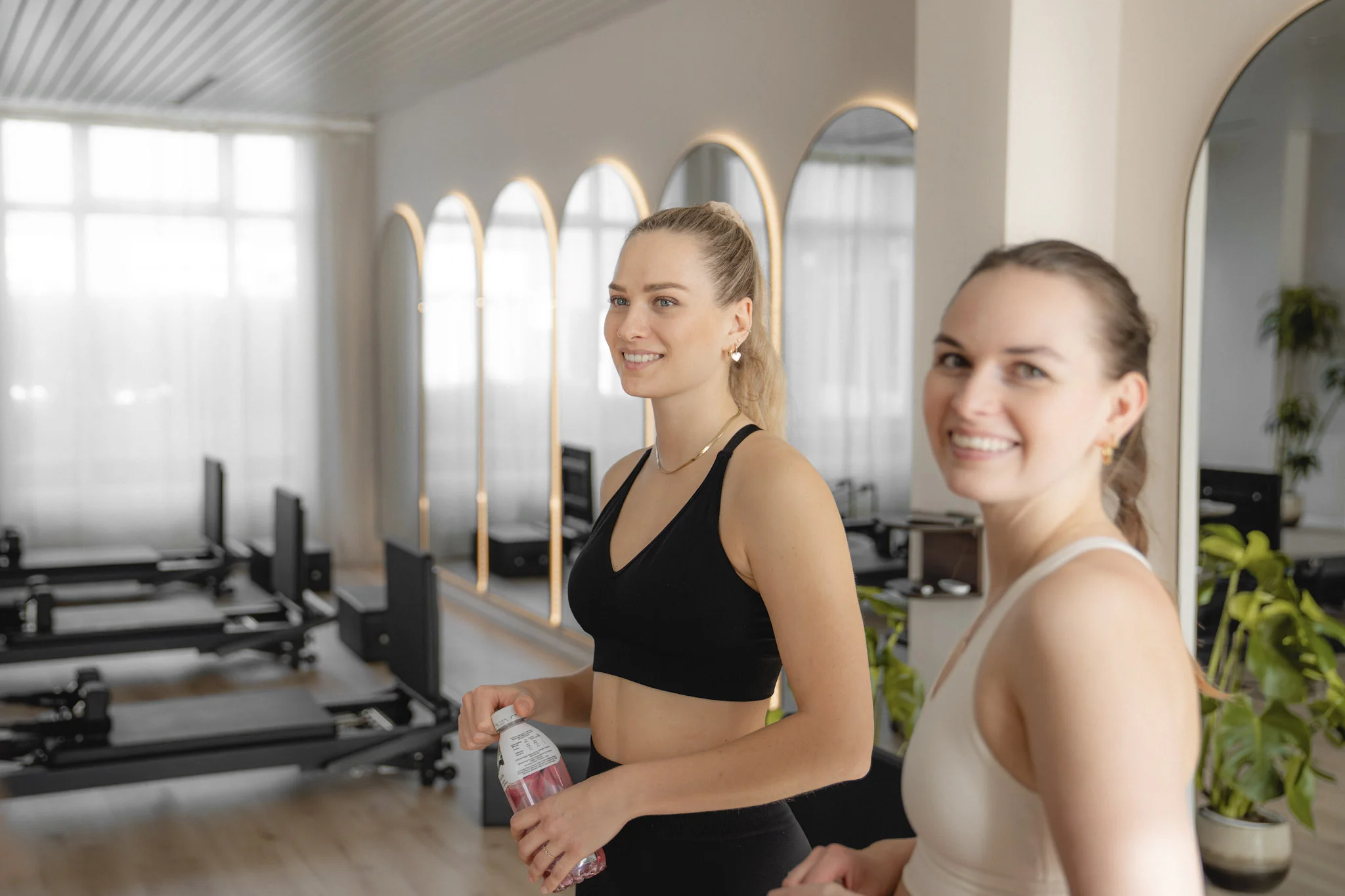Zwei lächelnde Frauen in Sportbekleidung in einem gut beleuchteten Pilates-Studio mit Spiegeln und Geräten im Hintergrund.