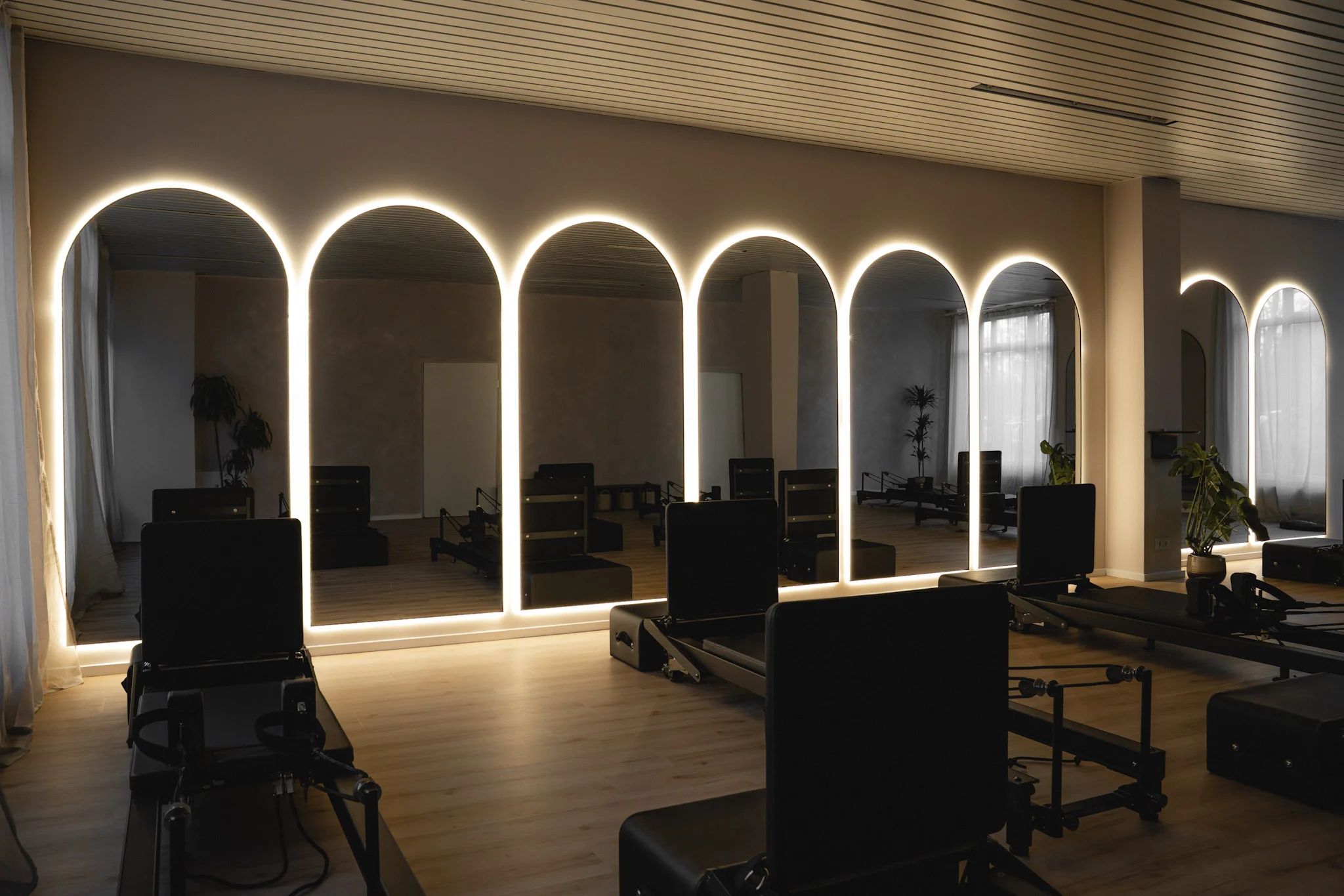 Moderner Fitnessraum mit Pilates-Geräten vor beleuchteten, bogenförmigen Spiegeln an der Wand.